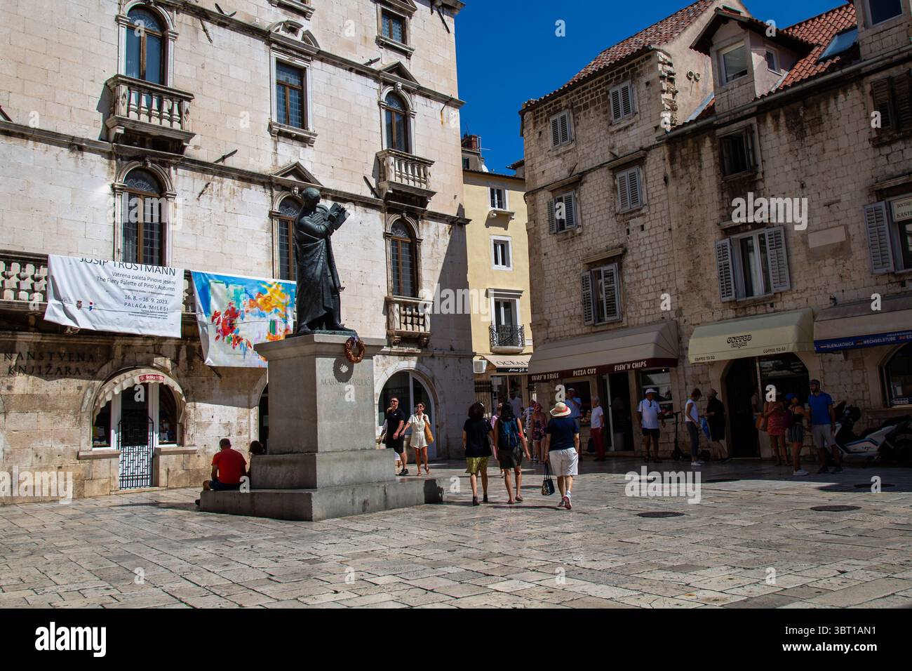 Spalato, Croazia - 8 settembre 2023: I turisti passeggiano in una piazza con una statua di Marko Marulić, vicino a negozi e una mostra d'arte. Foto Stock