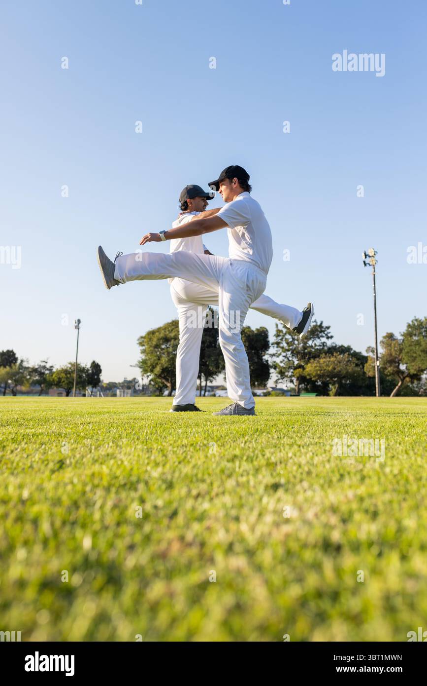 Diversi compagni di squadra maschili che si allungano in avanti sulle gambe sull'erba sotto il cielo limpido indossando uniformi bianche Foto Stock