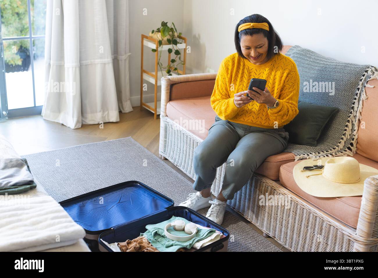 Donna rilassante che indossa un maglione giallo sul divano che controlla lo smartphone vicino a valigia aperta e spazio per le copie Foto Stock