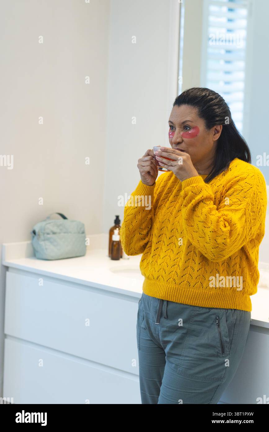 Donna matura con maglione giallo e toppe rosse sotto gli occhi sorseggiando dalla tazza al vanity, copia spazio Foto Stock