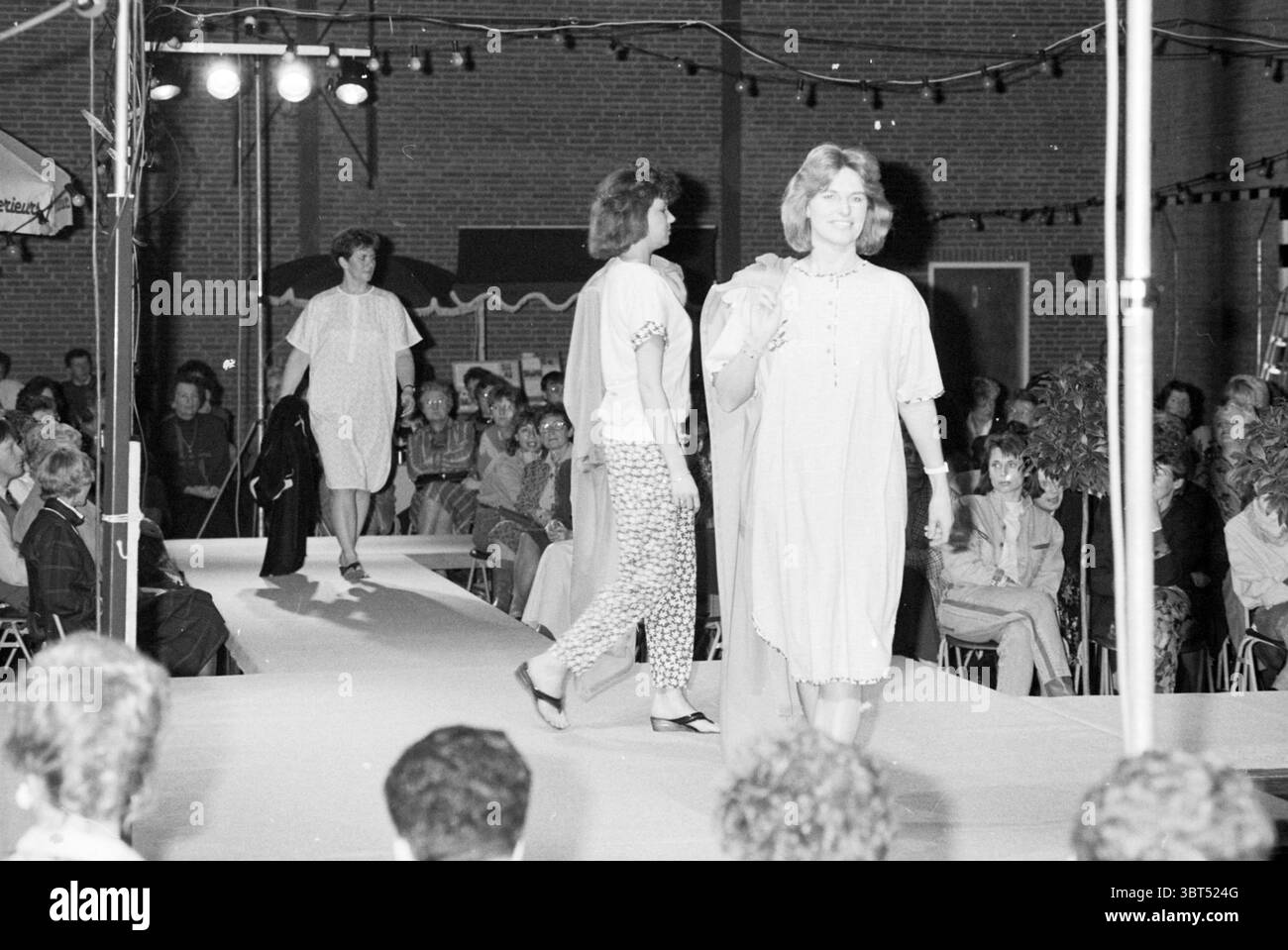 Sfilata di moda Spaarndam Fashion Spaarndam, Whizgle News, Dutch Desk, Paesi Bassi, 1950 - 2000 il 13-03-1989. L'immagine contiene questi argomenti. La scena si sviluppa in un ambiente di sfilata di moda, illuminato da luci di scena che creano un'atmosfera vibrante. In primo piano, un modello si muove lungo una passerella, indossando un abito aderente con un motivo sottile, completato da semplici sandali che consentono di muoversi facilmente. I suoi capelli sono modellati da onde morbide, incorniciando il suo volto e migliorando il suo comportamento sicuro mentre guarda davanti. Dietro di lei, un'altra modella si sta facendo strada lungo la passerella, dre Foto Stock