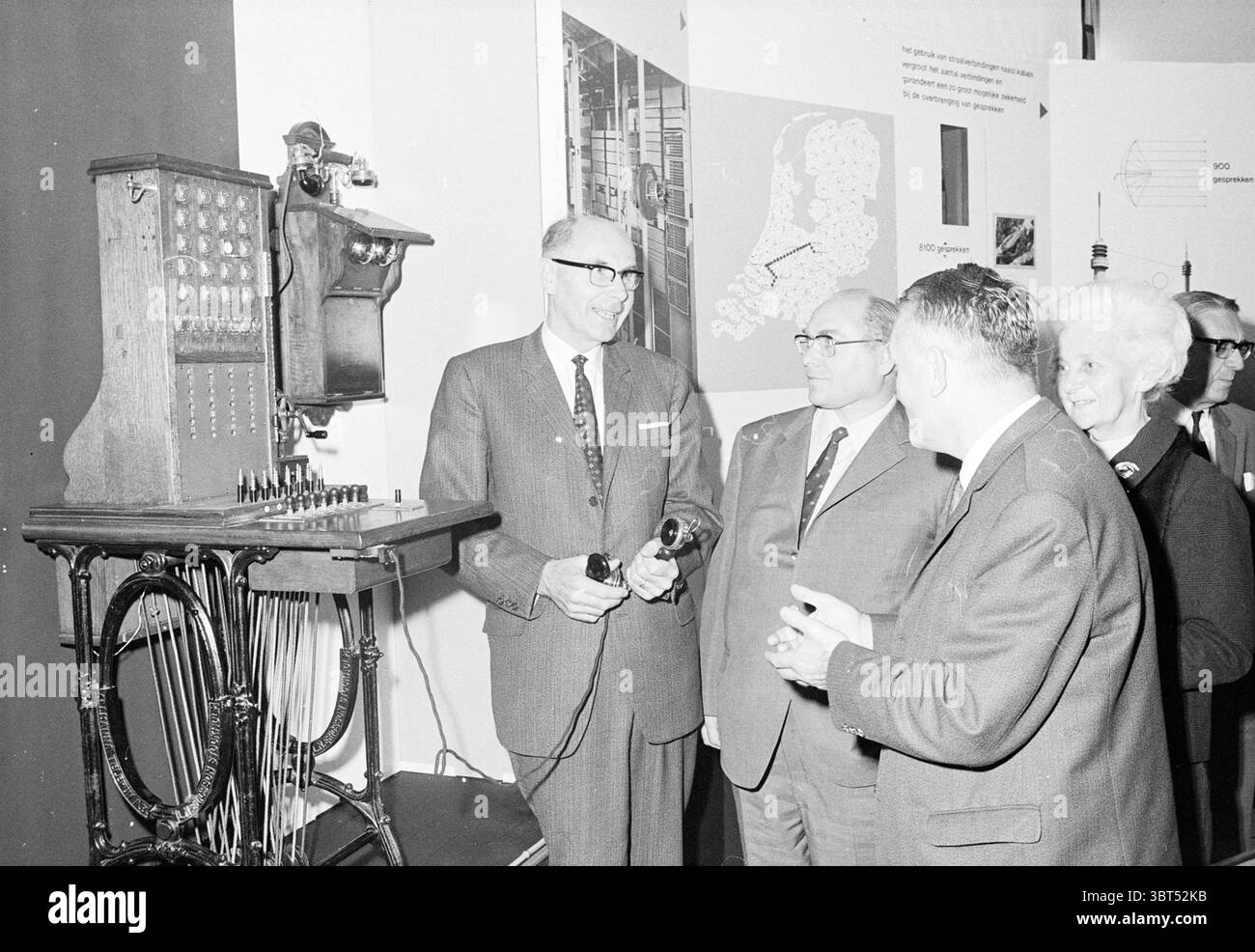 Apertura di una borsa telefonica apertura apertura apertura messa in servizio, Whizgle News, Dutch Desk, Paesi Bassi, 1950 - 2000 il 16-10-1970. L'immagine include questi argomenti. La scena cattura un raduno di diverse persone in un contesto espositivo storico. Al centro, due uomini sono impegnati in una conversazione, ognuno con i telefoni tradizionali. Indossano abiti formali, indicativi di uno stile della metà del XX secolo, con cravatte che riflettono toni tenui e motivi classici. Le loro espressioni facciali suggeriscono curiosità e interesse, magari discutendo della tecnologia di fronte a loro. Accanto al The Men, Foto Stock