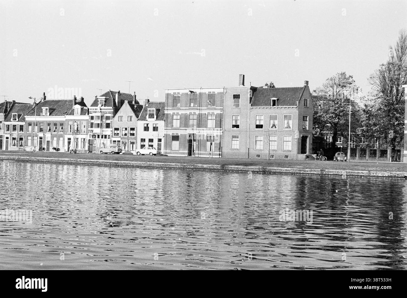 Leidsevaart Haarlem Leidsevaart Paesi Bassi., Whizgle News, Dutch Desk, Paesi Bassi, 1950 - 2000. Questi sono gli elementi dell'immagine. La scena raffigura una tranquilla vista sul fiume con una serie di edifici che costeggiano la riva. L'architettura presenta un mix di stili, con facciate tradizionali con tetti a spiovente, finestre strette e una varietà di finiture in mattoni e gesso. Le strutture sono dipinte in toni tenui, prevalentemente grigi e bianchi, con alcune case che mostrano colori pastello chiari che aggiungono un sottile calore alla composizione complessiva. Riflesso nell'acqua, negli edifici Foto Stock