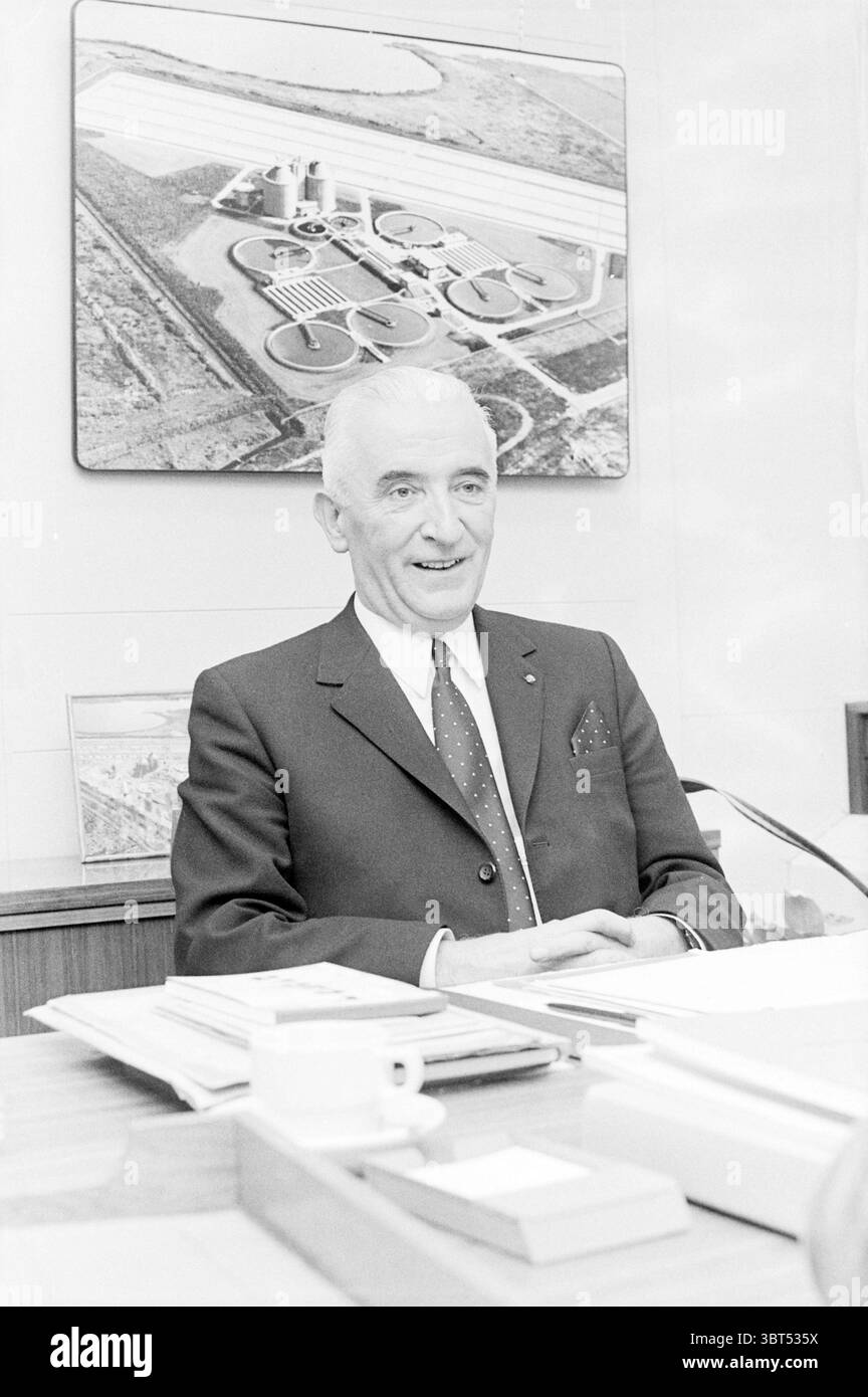 Ritratto IR. Fuykschot direttore tecnico dei lavori pubblici saluta Haarlem Paesi Bassi, Whizgle News, Dutch Desk, Paesi Bassi, 1950 - 2000 il 14-12-1971. Questi argomenti vengono visualizzati nell'immagine. Nella scena, un uomo sicuro di sé siede su una scrivania ordinata, caratterizzata da legno liscio e lucido. Indossa un abito scuro con una camicia bianca, completata da una cravatta di gusto e da una tasca quadrata che aggiunge un tocco raffinato. I suoi capelli sono curati e sfoggia un sorriso accogliente che trasmette calore e professionalità. Dietro di lui, una grande immagine incorniciata di una vista aerea di un comple Foto Stock