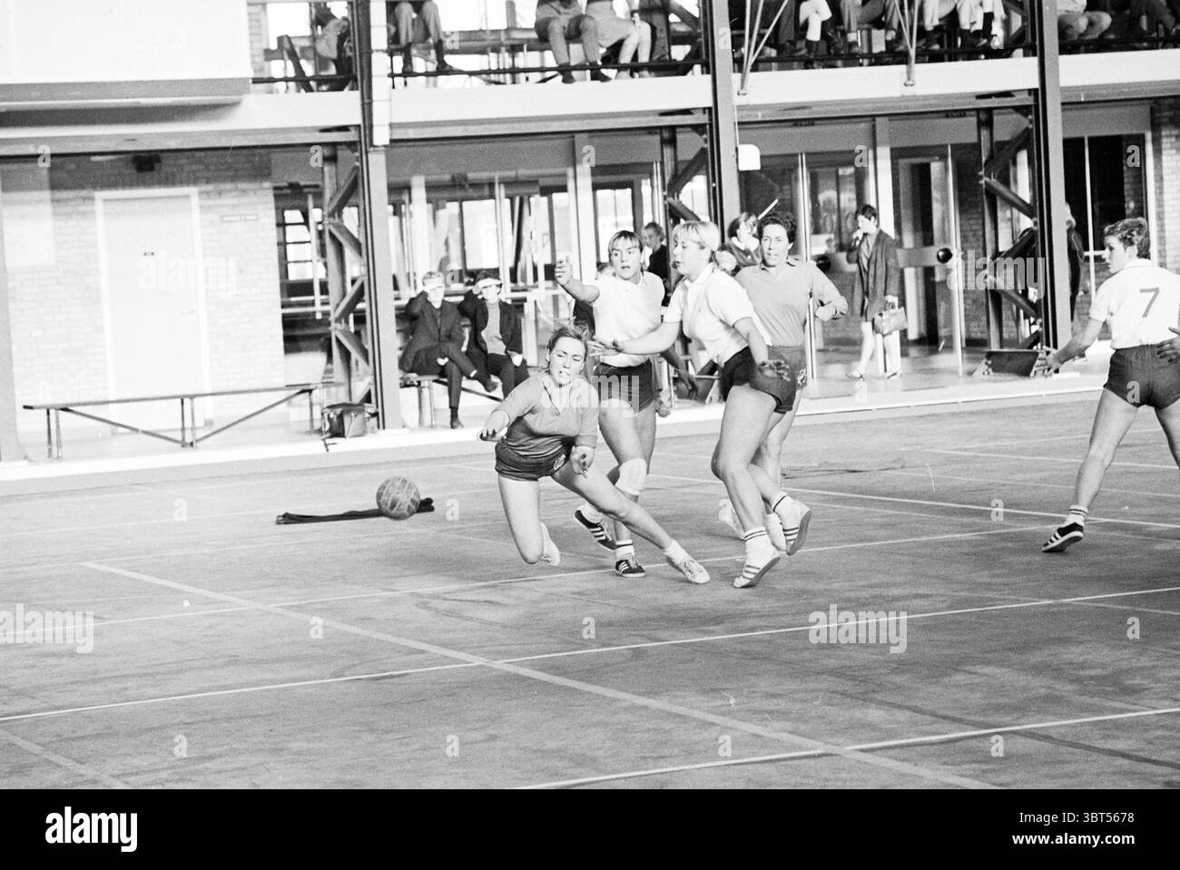 De Blinkert - Swiftde Blinkert - IJmond Handball, Whizgle News, Dutch Desk, Paesi Bassi, 1950 - 2000 il 10-12-1967. L'immagine mostra questi argomenti. In una struttura sportiva al coperto, una scena dinamica si svolge in uno scenario di architettura industriale. L'ambiente presenta un'area spaziosa con un pavimento liscio e scuro che contrasta con la luminosa illuminazione sopraelevata, creando un'atmosfera vivace. La tavolozza dei colori include tonalità di grigio e bianco, con tocchi di colore dalle uniformi degli atleti. Un gruppo di diversi individui, principalmente in abbigliamento sportivo, sono impegnati in un gioco vivace. Lo spettacolo Foto Stock