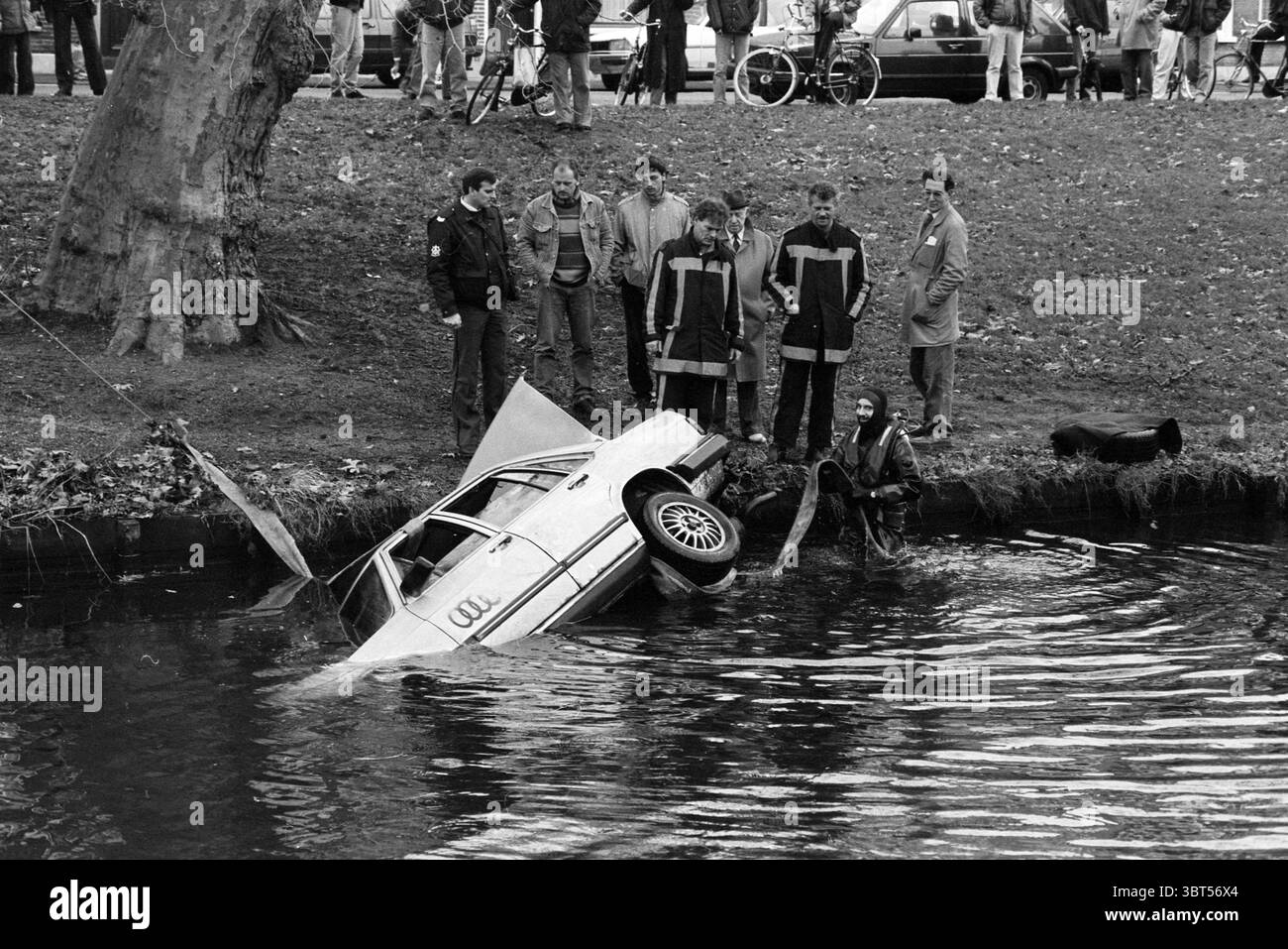 Car in water Raamsingel Haarlem Raamsingel Paesi Bassi, Whizgle News, Dutch Desk, Paesi Bassi, 1950 - 2000 il 20-12-1990. Questi argomenti sono mostrati nell'immagine. In una scena caratterizzata da un senso di urgenza, un veicolo parzialmente sommerso sporge dal bordo dell'acqua, con l'estremità posteriore profondamente immersa nelle profondità torbide. L'auto, un modello più vecchio, presenta un esterno bianco sbiadito, ora abbattuto dal tempo e dagli elementi. Intorno al veicolo, un gruppo di persone si trova su una riva erbosa, le cui espressioni sono un misto di curiosità e preoccupazione. La folla è vestita con una varietà di wi-fi oscuri e silenziosi Foto Stock