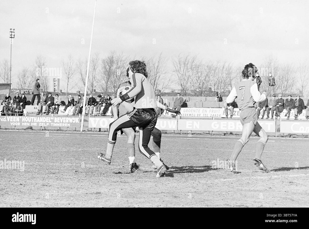 HBC - TYBB Football, Whizgle News, Dutch Desk, Paesi Bassi, 1950 - 2000 il 27-02-1977. Questi sono gli elementi dell'immagine. Una dinamica scena all'aperto si sviluppa su un campo erboso, dove l'intensità di una partita di calcio è palpabile. In primo piano, due giocatori sono impegnati nel caldo della competizione. Uno, rivestito in un'audace uniforme arancione, maneggia sapientemente la palla, mostrando abilità in movimento. Il giocatore avversario, vestito con una divisa rossa adornata con il numero sette, si prepara a fare una manovra difensiva. Lo sfondo rivela un raduno di spettatori seduti sulle gradinate, creando un mosaico di va Foto Stock