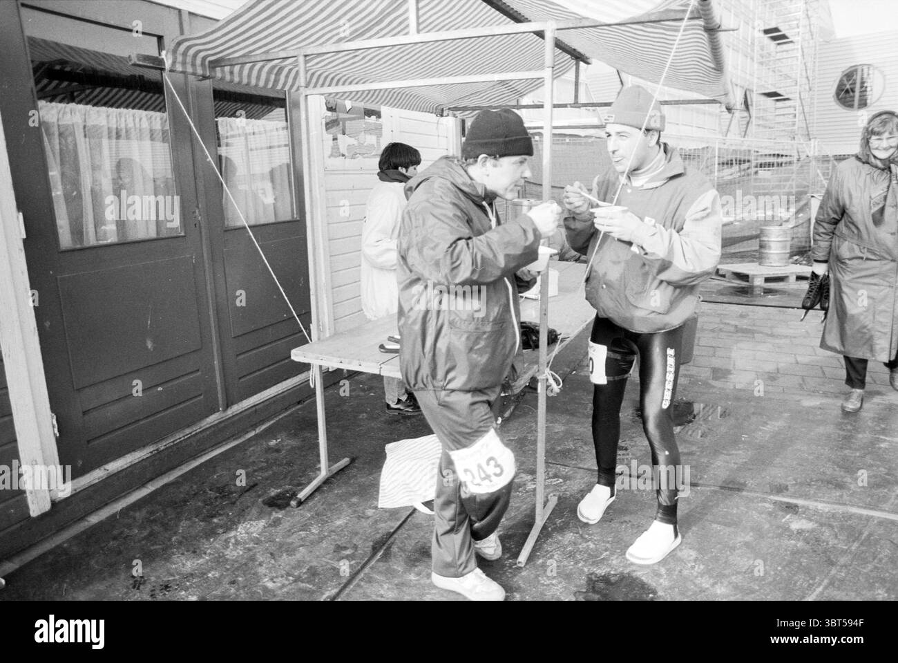 Triathlon invernale. Haarlem Haarlem Paesi Bassi, Whizgle News, Dutch Desk, Paesi Bassi, 1950 - 2000 il 19-02-1993. L'immagine include questi argomenti. In una vivace scena all'aperto, due persone vestite per il freddo si stanno impegnando in una conversazione vicino a un rifugio temporaneo. Il rifugio è contrassegnato da una tettoia a strisce, che fornisce un piccolo rifugio dal freddo. Una persona che indossa un berretto scuro e un cappotto lungo tiene una piccola tazza, apparentemente bevendo una bevanda calda. Ha un piccolo numero fissato al suo cappotto, suggerendo la partecipazione a un evento. L'altra persona, vestita con un outfit atletico aderente Foto Stock