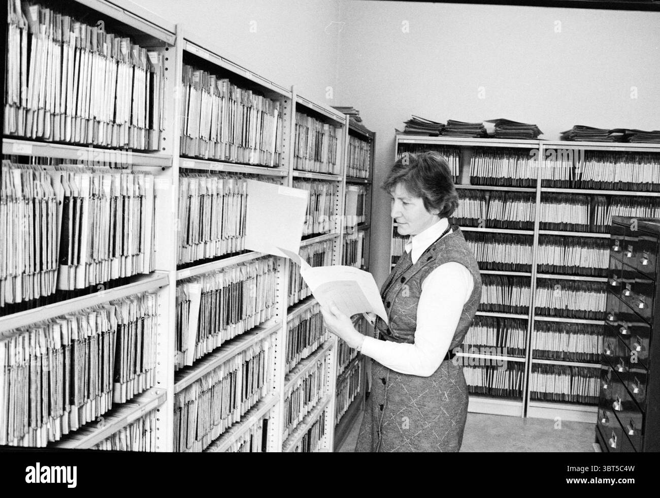 Donna al deposito di alloggi, Whizgle News, Dutch Desk, Paesi Bassi, 1950 - 2000 il 11-04-1979. Questi sono gli elementi dell'immagine. La scena cattura una donna in piedi in una stanza piena di scaffali alti ricchi di cartelle e documenti ordinati. Gli scaffali, che si estendono dal pavimento al soffitto, sono pieni di lime che sono principalmente in tonalità di beige e grigio, conferendo allo spazio un aspetto monocromatico. La donna è vestita con una blusa di colore chiaro posta sotto un gilet scuro e testurizzato, creando un contrasto con il top. I suoi capelli sono modellati con onde morbide e lei sembra concentrata come Foto Stock