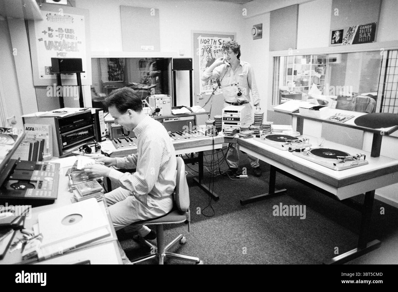 Persone nello studio di RTV Noord-Holland, Whizgle News, Dutch Desk, Paesi Bassi, 1950 - 2000 il 04-09-1990. L'immagine mostra questi argomenti. La scena è ambientata in un vivace studio radio, pieno di un eclettico mix di attrezzature e arredi che mette in risalto un senso di creatività e nostalgia. A sinistra, una persona è seduta a una scrivania, concentrandosi su una serie di apparecchiature audio, tra cui un'ampia console di missaggio, un deck per cassette e dischi in vinile sparsi intorno. Sono vestiti in modo informale, suggerendo un'atmosfera rilassata ma impegnata. L'artista è in profonda concentrazione, con le mani che agiscono Foto Stock