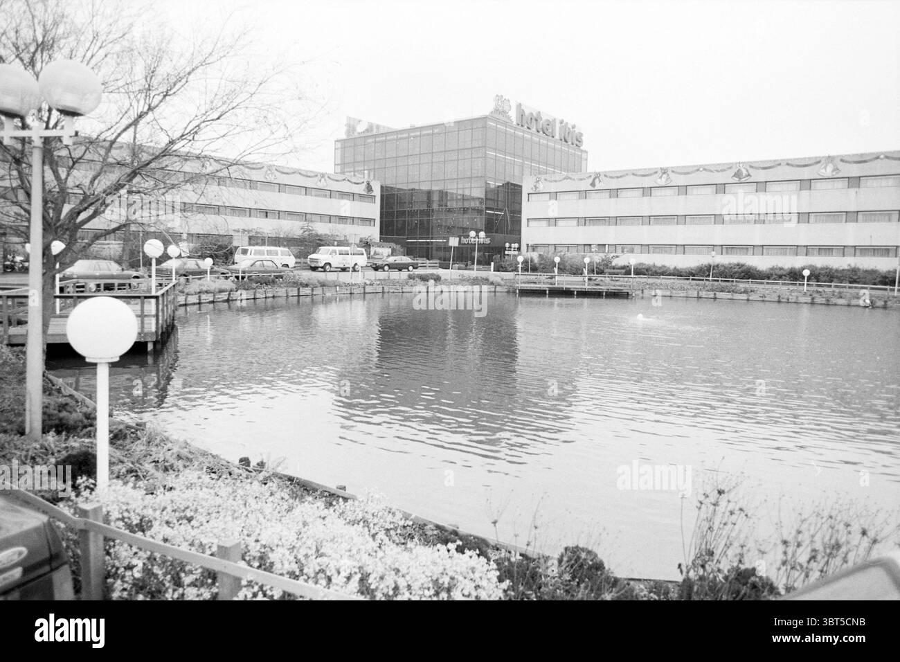 Hotel Ibis Outside Hotels, Whizgle News, Dutch Desk, Paesi Bassi, 1950 - 2000 on 08-12-1988. Questi argomenti sono mostrati nell'immagine. La scena cattura una giornata serena e leggermente ricoperta in un ambiente urbano. In primo piano, un corpo d'acqua calmo e riflettente forma un piccolo laghetto, circondato da un'area ben curata con vari arbusti e piante basse, suggerendo un'attenta manutenzione. Un gruppo di lampade sferiche bianche si erge su pali lungo il bordo dell'acqua, creando un'atmosfera morbida e invitante. Oltre il laghetto, un moderno edificio a più piani e' prominente, che presenta grandi finestre in vetro che mirro Foto Stock