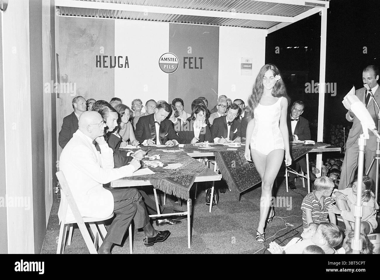 Miss Coast elezioni con Albert mol e Lou van Rees nella giuria, Whizgle News, Dutch Desk, Paesi Bassi, 1950 - 2000 il 06-08-1967. L'immagine include questi argomenti. La scena cattura un vivace evento all'aperto, probabilmente una sfilata di moda o un concorso, ambientato su uno sfondo caratterizzato da una combinazione di colori audaci. Un palcoscenico luminoso con sezioni rosse e bianche attira l'attenzione, contrastando con l'ambiente scuro, suggerendo un ambiente serale. In primo piano, un modello si muove in tutta sicurezza lungo una passerella, mostrando un elegante outfit bianco dalla forma aderente. La sua postura trasmette il suo equilibrio e l'eleganza, con Foto Stock