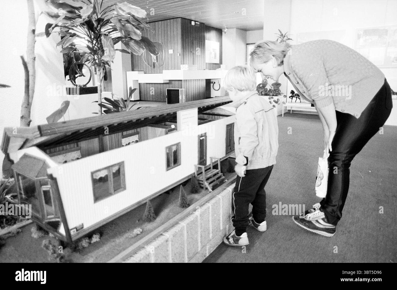 Servizio di pianificazione provinciale allestimento di mostre e residenti carovane mostre carovane campi carovanieri residenti caravan, Whizgle News, Dutch Desk, Paesi Bassi, 1950 - 2000 il 12-08-1986. Questi sono gli elementi dell'immagine. La scena cattura una calda interazione tra un adulto e un bambino, entrambi concentrati su un modello dettagliato di una casa. Il modello ha un design pulito e moderno, caratterizzato da un esterno bianco con accenti scuri contrastanti. L'adulta, indossata con un maglione grigio chiaro, si inclina leggermente verso il basso, mostrando un atteggiamento nutriente mentre osserva attentamente il modello accanto al c Foto Stock