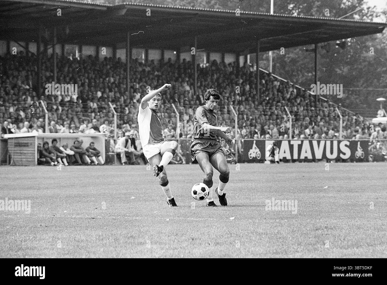 Haarlem - Ajax Football Haarlem, Whizgle News, Dutch Desk, Paesi Bassi, 1950 - 2000 il 14-08-1977. L'immagine contiene questi argomenti. La scena si svolge su un vivace campo da calcio, dove due giocatori sono impegnati dinamicamente in una partita. A sinistra, un giocatore con un'uniforme di colore chiaro sta eseguendo un potente calcio, una gamba sollevata in alto, catturando l'intensità dell'azione. La sua espressione riflette la determinazione, sottolineando lo spirito competitivo del gioco. Di fronte a lui, un altro giocatore rivestito di un'uniforme più scura è pronto, concentrandosi sul pallone da calcio ai suoi piedi. Il campo è circondato da un grande cro Foto Stock