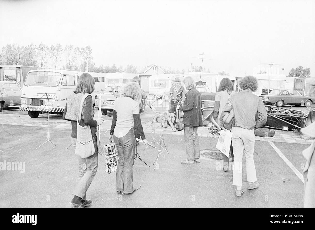 Il Ricciotti Ensemble è un'orchestra sinfonica di strada olandese? Esibizione in un parcheggio per roulotte., Whizgle News, Dutch Desk, Paesi Bassi, 1950 - 2000. L'immagine mostra questi argomenti. La scena cattura un gruppo di individui, prevalentemente giovani adulti, che camminano insieme attraverso un'area pavimentata che somiglia a un parcheggio. L'atmosfera è rilassata e ha un'aria di nostalgia, suggerendo un incontro informale o un evento. In primo piano, molte persone sono vestite con stili che ricordano la fine degli anni '1960 e l'inizio degli anni '1970, tra cui jeans con fondo a campana, camicie a motivi geometrici e gilet. I loro capelli sono lunghi e naturali Foto Stock