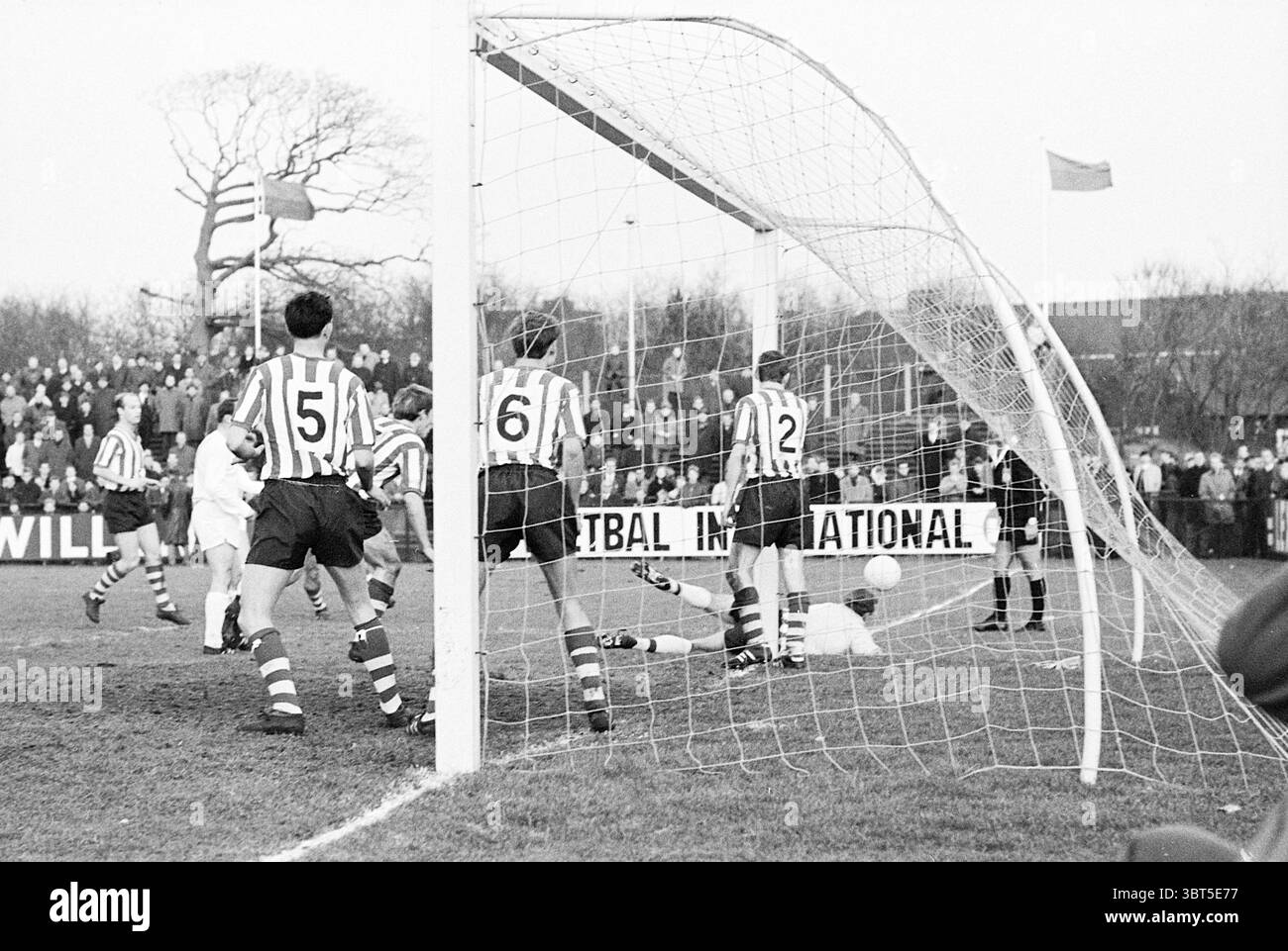 Partita di calcio., Whizgle News, Dutch Desk, Paesi Bassi, 1950 - 2000. Questi sono gli argomenti nell'immagine. La scena cattura un momento dinamico in una partita di calcio ambientata in quello che sembra essere uno stadio locale. Il primo piano è caratterizzato da un goalpost, la sua cornice bianca leggermente angolata, suggerendo un gioco vigoroso. La rete, allungata e leggermente sporgente, indica un recente tentativo di punteggio. Sullo sfondo, i giocatori vestiti di maglie a strisce, con tonalità alternate di rosso e bianco, sono impegnati nel caldo della competizione. Il loro obiettivo è diretto verso il pallone, che è probabilmente vicino al portiere. L'obiettivo Foto Stock