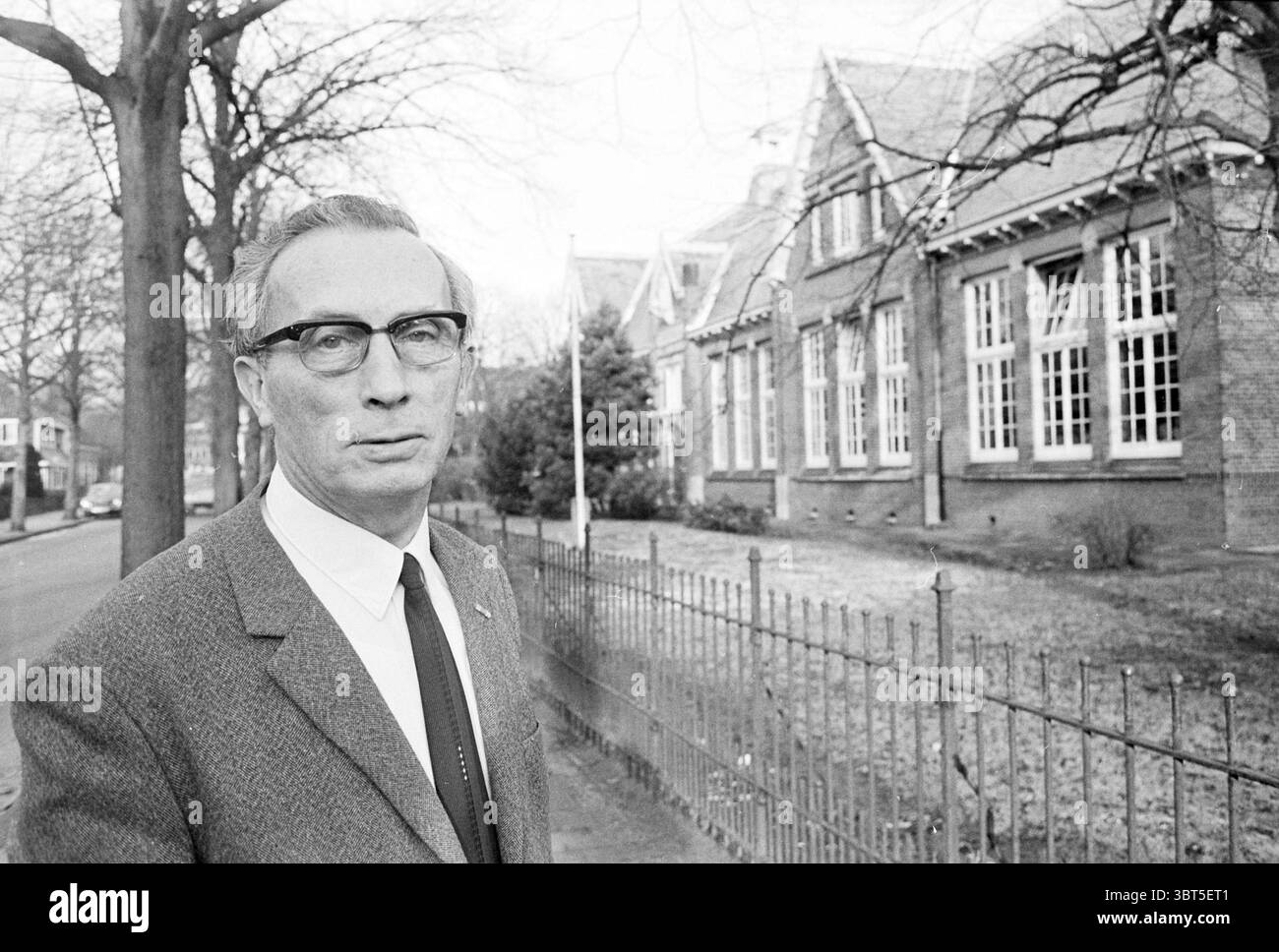 Ritratto del capo della St Joseph School Schools, Whizgle News, Dutch Desk, Paesi Bassi, 1950 - 2000 il 19-11-1971. L'immagine contiene questi argomenti. In un ambiente urbano sereno, un uomo distinto si trova in primo piano, trasudando un senso di quiete autorità. Indossa un abito sartoriale grigio scuro impreziosito da una camicia bianca nitida e una cravatta stretta, il tutto per aggiungere un aspetto sofisticato. La sua acconciatura è ben pettinata e sfoggia occhiali sottili e rettangolari che sottolineano il suo comportamento premuroso. Dietro di lui, un edificio in mattoni presenta grandi finestre a più pannelli, che riflettono Foto Stock