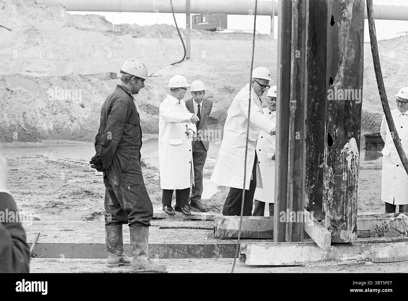 1st post PEN Central primo post, Whizgle News, Dutch Desk, Paesi Bassi, 1950 - 2000 on 26-05-1971. Questi argomenti sono mostrati nell'immagine. La scena raffigura un cantiere o un sito industriale, caratterizzato da un mix di attività e osservazione. In primo piano, un lavoratore vestito con abiti scuri e robusti e stivali pesanti ha un'espressione seria, osservando il processo in corso. Indossa un elmetto, segnalando le precauzioni di sicurezza in un ambiente potenzialmente pericoloso. Nelle vicinanze, un gruppo di individui è riunito intorno a un grande apparato simile a un trapano. Queste cifre sono vestite di colore chiaro Foto Stock