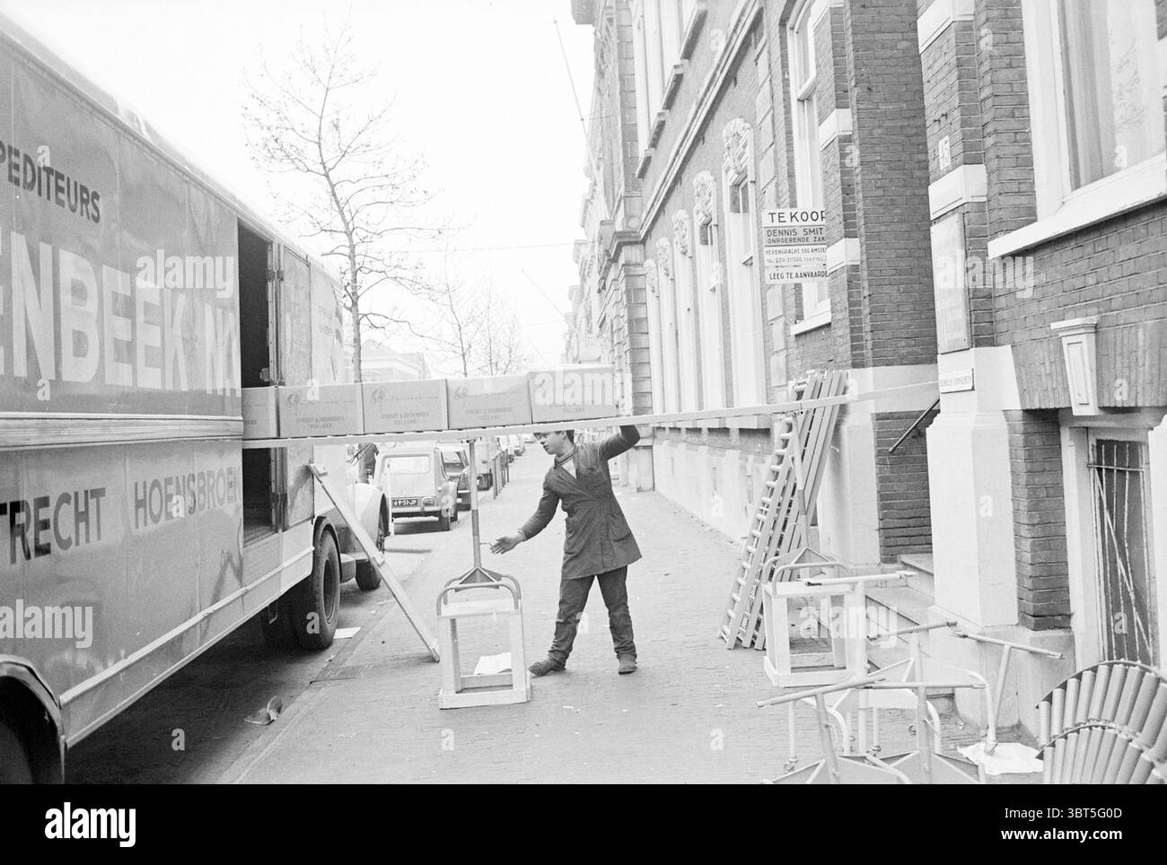 Trasferimento della cultura dei bulbi di fiori, Whizgle News, Dutch Desk, Paesi Bassi, 1950 - 2000 il 14-04-1971. L'immagine mostra questi argomenti. La scena raffigura una vivace strada urbana, caratterizzata da uno schema di colori monocromatico che enfatizza le sfumature di grigio. Un autocarro per consegne si trova in posizione prominente sulla sinistra, con il lato aperto che rivela le casse di legno scaricate. Le casse sono di colore terroso, in contrasto con la superficie metallica del veicolo. In primo piano, un uomo, vestito con un cappotto scuro e un cappello, è attivamente impegnato nel sollevare una delle casse. La sua postura suggerisce determinazione e. Foto Stock