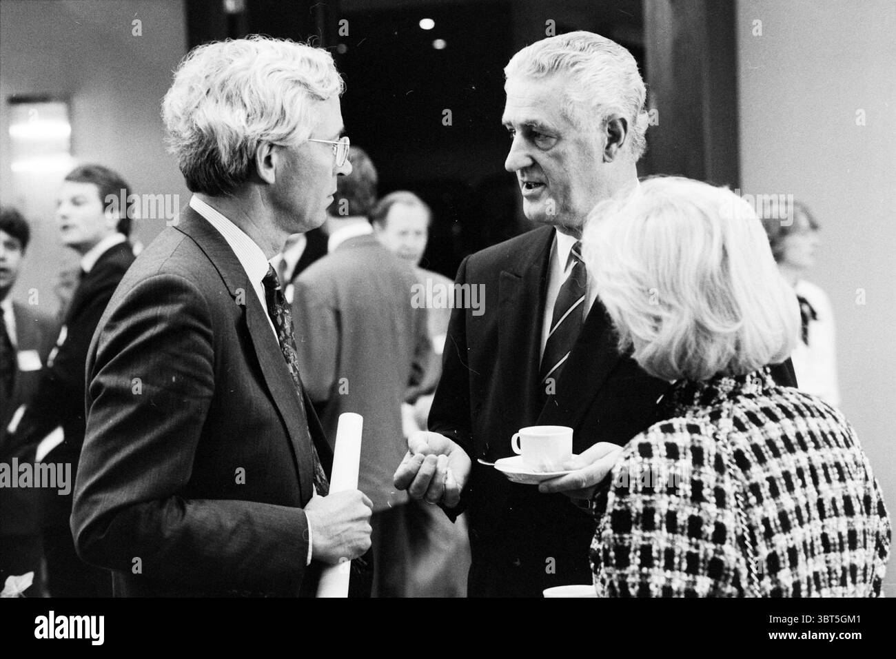 M. Schröder Schiphol Hilton Chamber of Commerce Schiphol, Whizgle News, Dutch Desk, Paesi Bassi, 1950 - 2000 il 22-04-1991. L'immagine mostra questi argomenti. In un vivace ambiente interno, un gruppo di persone si impegna a conversare, caratterizzato da un mix di abbigliamento formale e informale. Due uomini sono prominenti in primo piano; entrambi hanno un aspetto distinto, con un uomo che sfoggia capelli argentati e indossa un abito scuro, mentre l'altro ha capelli di colore chiaro disegnati con cura ed è vestito con un abito grigio. L'uomo dai capelli d'argento tiene una tazza in una mano, gestendo con l'altra mentre parla, h Foto Stock