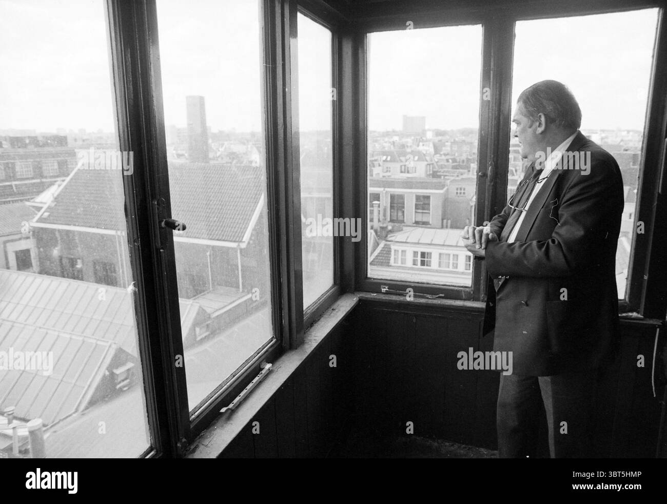 Tour guidato di Enschede, Whizgle News, Dutch Desk, Paesi Bassi, 1950 - 2000 il 23-05-1991. Questi argomenti vengono visualizzati nell'immagine. La scena cattura un uomo in piedi davanti a una grande finestra in un alto punto panoramico, che si affaccia su un paesaggio urbano. È vestito con un abito scuro, che contrasta con l'atmosfera più luminosa all'esterno. La sua postura suggerisce la contemplazione mentre guarda fuori, le mani si stringono insieme, forse perse nel pensiero. La finestra incornicia una vista che rivela un mix di stili architettonici, con tetti di vari design, alcuni piatti e altri picchi, creando uno skyline eclettico. Gli edifici sono un buio Foto Stock