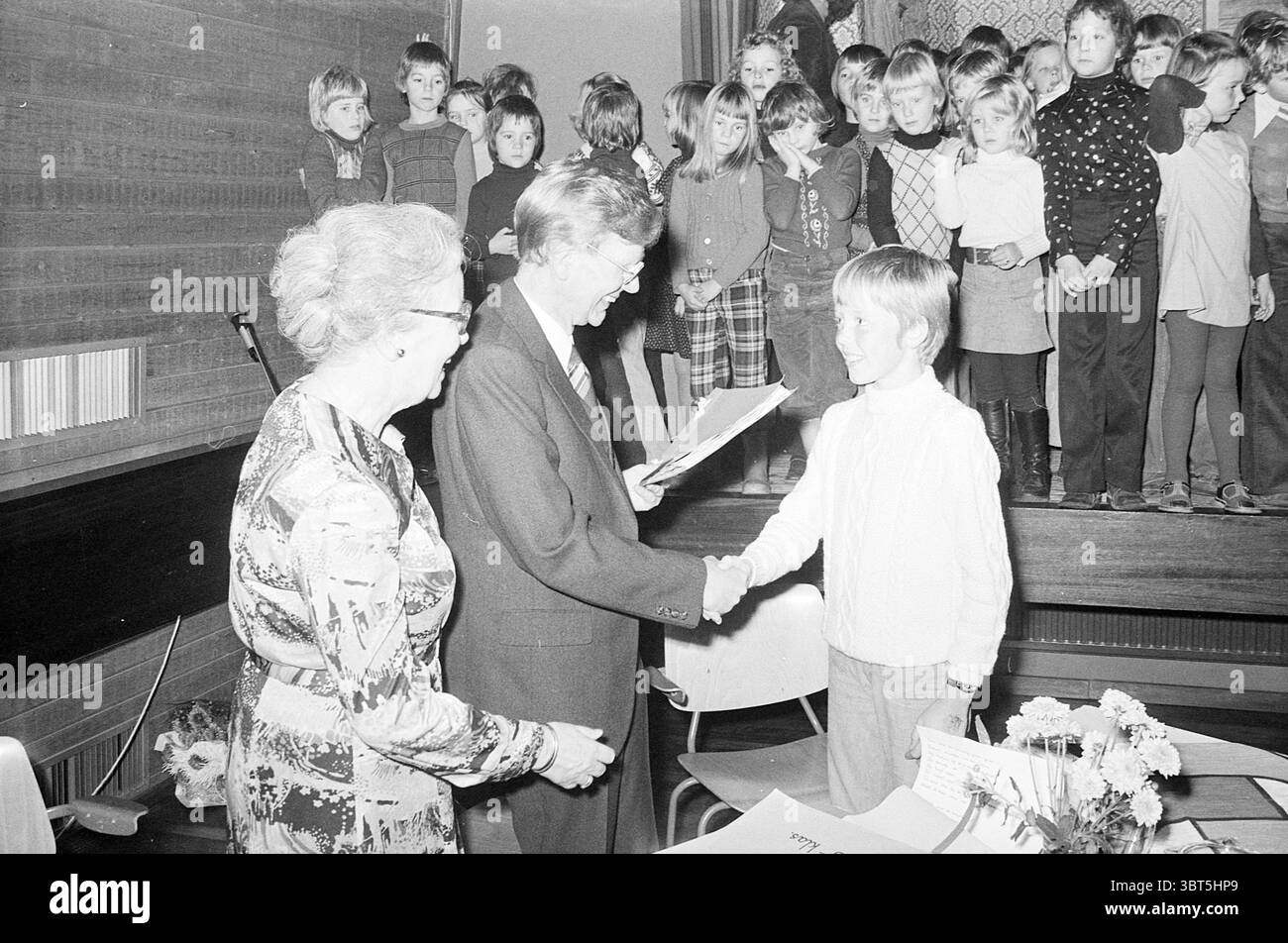 Uomo donna e ragazzo con bambini sul palco sullo sfondo., Whizgle News, Dutch Desk, Paesi Bassi, 1950 - 2000. L'immagine include questi argomenti. In un ambiente caldo e nostalgico, un gruppo di bambini si erge su una piattaforma rialzata sullo sfondo, tutti vestiti con un mix di maglioni colorati e abiti a fantasia, irradiando innocenza e gioia. Le loro espressioni vanno dall'anticipazione all'eccitazione, indicando un momento di festa. I bambini sono strettamente raggruppati, creando un senso di comunità e un'esperienza condivisa. In primo piano, due adulti si impegnano in un momento di riconoscimento o. Foto Stock