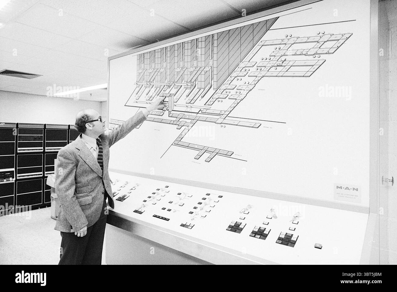 Magazzino MSD + computer Haarlem Buildings, Whizgle News, Dutch Desk, Paesi Bassi, 1950 - 2000 il 07-10-1977. L'immagine include questi argomenti. La scena è ambientata in una sala grande e funzionale con un ambiente moderno ma industriale. Un uomo si erge a sinistra, vestito con una tuta grigia formale con una distinta cravatta a motivi geometrici, che aggiunge un tocco di raffinatezza all'ambiente. Sta puntando verso un grande diagramma tecnico montato sul muro, che delinea un layout complesso, probabilmente correlato a macchinari o sistemi informatici. Il diagramma è prevalentemente bianco con varie linee e schemi Foto Stock