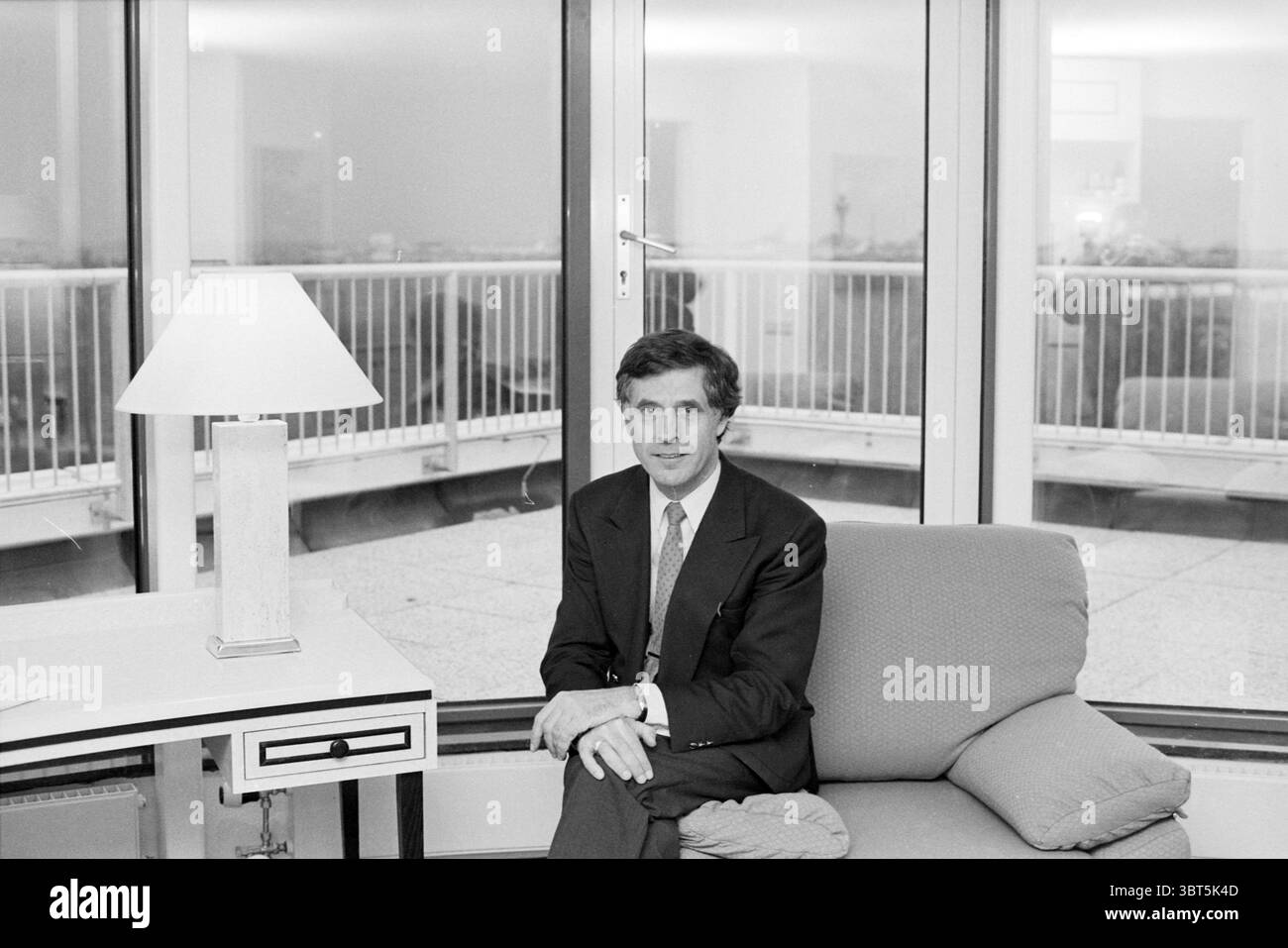 Nooijens Dorint hotel Haarlemmermeer, Whizgle News, Dutch Desk, Paesi Bassi, 1950 - 2000 il 12-11-1991. Questi sono gli argomenti nell'immagine. La scena è ambientata in un moderno spazio interno, caratterizzato da grandi finestre in vetro che consentono alla luce naturale di filtrare, creando un'atmosfera morbida e invitante. Le pareti sono probabilmente di colore chiaro, contribuendo a creare un'atmosfera pulita e sofisticata. In primo piano, un individuo ben vestito siede comodamente su un divano di colore chiaro che appare lussuoso e invitante. Il suo abbigliamento è formale e presenta un abito sartoriale che contrasta con l'eleganza casual del t Foto Stock