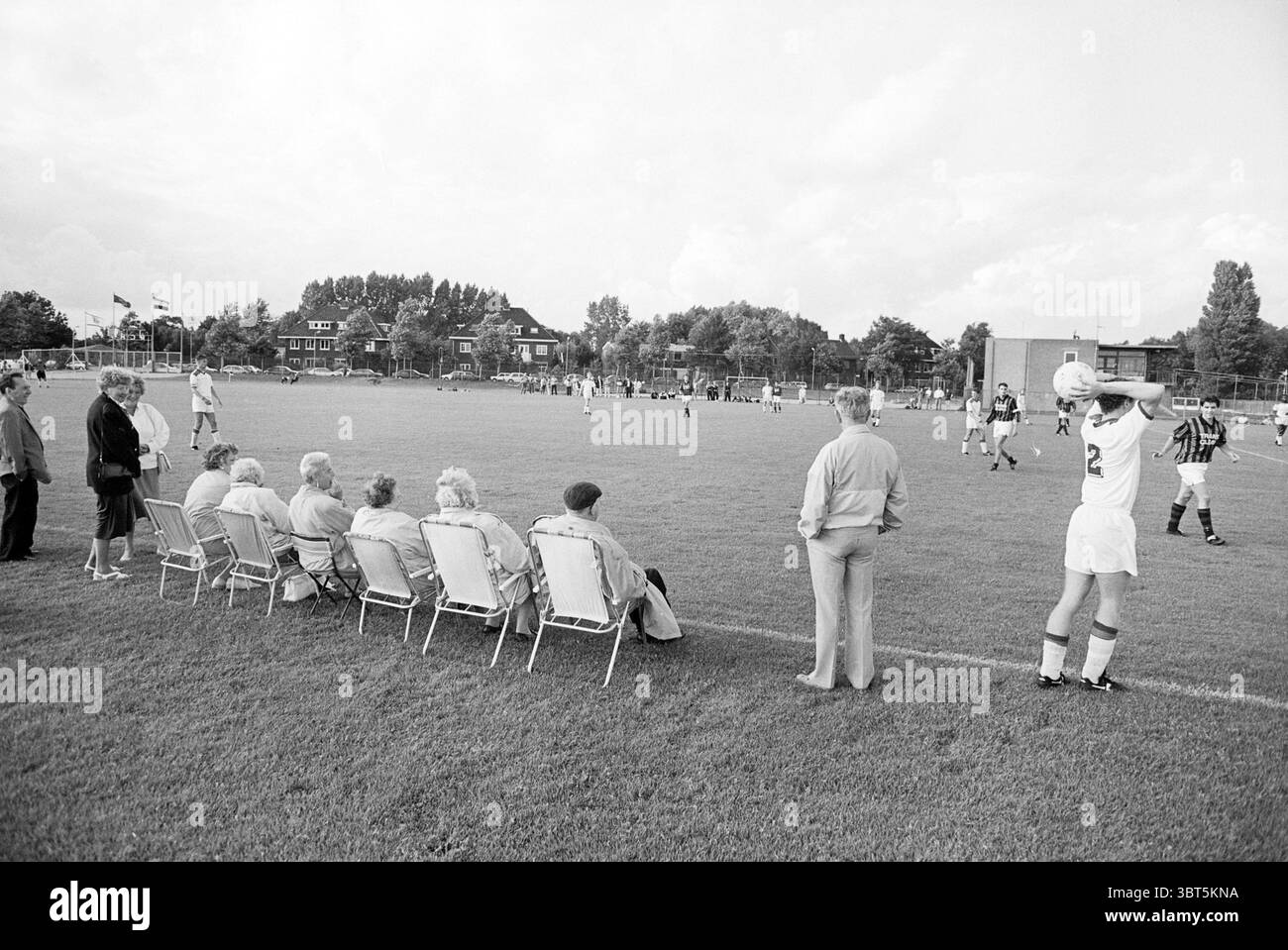 Telstar 2 - RCH Haarlem Cup, Whizgle News, Dutch Desk, Paesi Bassi, 1950 - 2000 il 30-07-1993. L'immagine mostra questi argomenti. La scena raffigura un vivace campo da calcio all'aperto. In primo piano, un gruppo di spettatori siede su una fila di sedie pieghevoli in metallo, osservando il gioco con diverse espressioni di concentrazione e divertimento. Sono vestiti con giacche e pantaloni dai colori chiari, che suggeriscono un abbigliamento casual ma premuroso per un'escursione. Oltre a loro, la lussureggiante erba verde del campo contrasta nettamente con il cielo grigio e coperto, suggerendo un imminente cambiamento del tempo. Il lettore Foto Stock