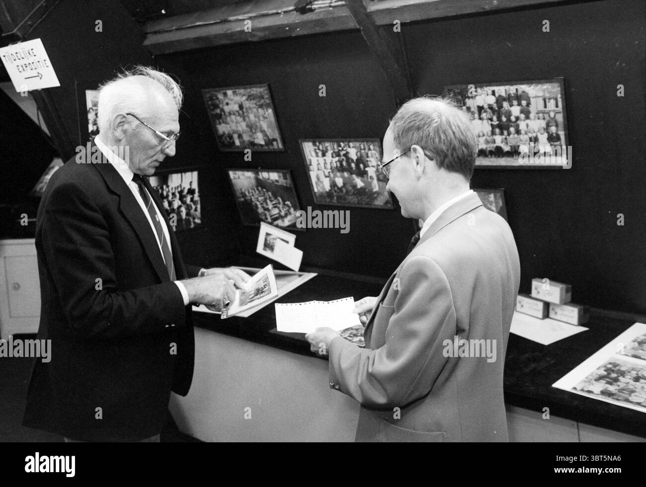 2 fondatori del museo Bavo Noordwijkerhout Museum Museums Noordwijkerhout, Whizgle News, Dutch Desk, Paesi Bassi, 1950 - 2000 il 28-05-1986. Questi sono gli argomenti nell'immagine. La scena presenta due persone coinvolte in una conversazione in uno spazio espositivo o espositivo. Si trovano di fronte a un lungo bancone scuro, sormontato da una varietà di fotografie disposte in una griglia su sfondo nero. L'illuminazione è soffusa e produce ombre sottili che contribuiscono a creare un'atmosfera calda e silenziosa. A sinistra, un uomo anziano con capelli e occhiali grigi indossa un abito scuro con una camicia di colore chiaro. Lui Foto Stock