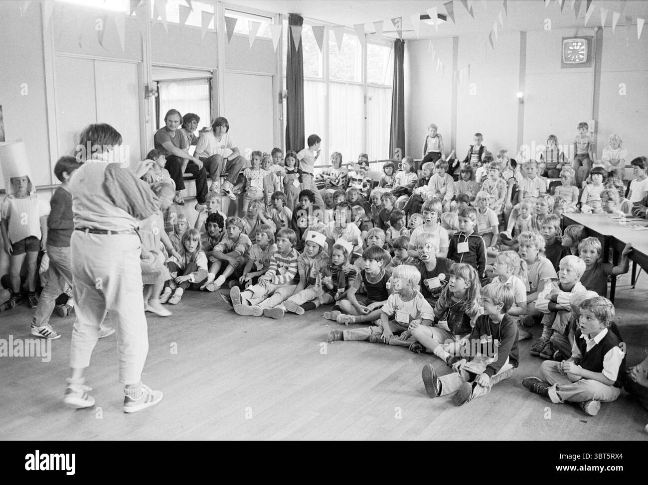 I figli di Doedag, Joh. parish Hillegom Children's Homes Children's Party Children's Games and toys Hillegom, Whizgle News, Dutch Desk, Paesi Bassi, 1950 - 2000 il 17-07-1984. L'immagine include questi argomenti. La scena si svolge in una sala spaziosa e ben illuminata, decorata con coloratissimi striscioni triangolari appesi al soffitto. Le pareti sono dipinte in toni delicati e neutri, aggiungendo un'atmosfera allegra ma rilassante. Un grande gruppo di bambini riempie il primo piano, seduti ordinatamente sul pavimento, i loro volti adornati da espressioni di curiosità ed eccitazione. Sono vestiti in una varietà Foto Stock
