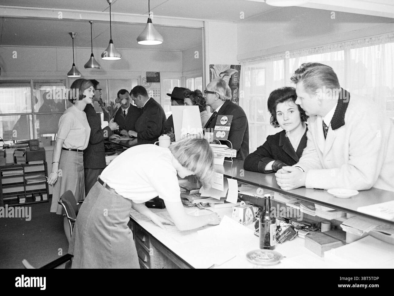 Personale e clienti al banco di un'agenzia di viaggi 1960S.. Ufficio turistico., Whizgle News, Dutch Desk, Paesi Bassi, 1950 - 2000. Questi argomenti vengono visualizzati nell'immagine. In un ambiente d'ufficio vivace, un gruppo di professionisti si impegna in varie attività. L'impostazione è resa più luminosa dalla luce soffusa e ambientale prodotta da più apparecchi sospesi, che proietta un caldo bagliore sulla scena. A sinistra, una donna vestita elegantemente parla animatamente con un'altra persona, le sue espressioni suggeriscono una conversazione vivace. Entrambi sono circondati da colleghi, alcuni dei quali si fermano nelle vicinanze, che condividono le discussioni. Foto Stock