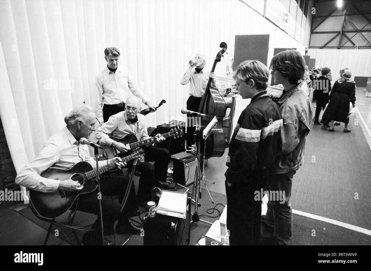Spring Fair senior orchestra, Whizgle News, Dutch Desk, Paesi Bassi, 1950 - 2000 il 28-03-1991. Questi argomenti sono mostrati nell'immagine. In uno spazioso ambiente interno con alti soffitti e uno sfondo foderato, si svolge un raduno che irradia un'atmosfera vivace ma intima. Un gruppo di uomini più anziani è impegnato in una performance musicale, ognuno profondamente concentrato sui propri strumenti. Un individuo, seduto in modo prominente, suona una chitarra acustica, mentre un altro toglie le corde di un contrabbasso in piedi nelle vicinanze. Le loro espressioni trasmettono piacere e concentrazione, riflettendo una passione condivisa per la musica. Su Foto Stock