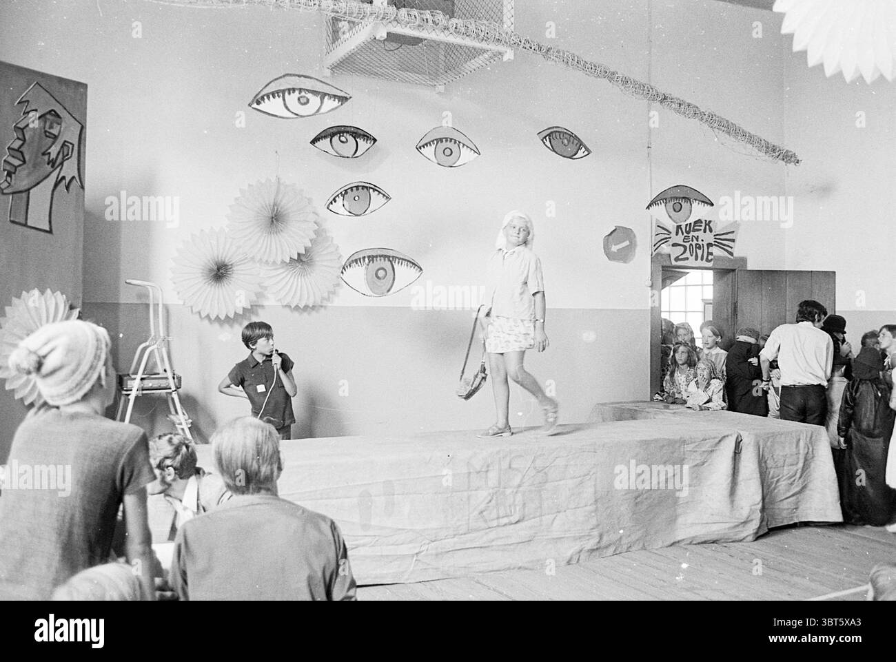 Elezione Miss Krat Theater, Whizgle News, Dutch Desk, Paesi Bassi, 1950 - 2000 il 09-08-1969. Questi argomenti sono mostrati nell'immagine. In un ambiente interno spazioso, un'atmosfera vibrante è creata da una serie di grandi occhi astratti appesi alla parete, donando allo spazio un tocco artistico. Questi occhi, disegnati con contorni audaci, contrastano nettamente con i toni più morbidi della stanza. Al centro, una piattaforma rialzata funge da punto focale per un'esposizione di moda. Una giovane modella corre con fiducia lungo questa passerella, indossando un abito leggero e fantasia che cattura l'attenzione e riflette un po' di divertimento Foto Stock