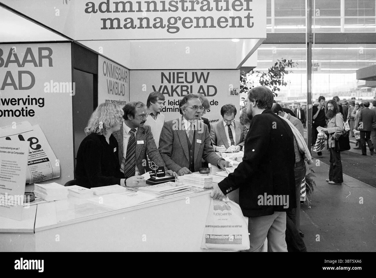 Gli editori di Diligentia sono presenti a una fiera, Whizgle News, Dutch Desk, Paesi Bassi, 1950 - 2000 il 21-10-1980. Questi argomenti sono mostrati nell'immagine. L'ambiente è un vivace spazio per eventi, probabilmente un convegno o una fiera, caratterizzato da un soffitto alto e un ampio piano aperto. Le luci fluorescenti creano un'illuminazione brillante e uniforme, contribuendo a creare un'atmosfera vivace e laboriosa. Uno stand di spicco cattura l'attenzione con il suo design elegante e moderno. Presenta una delicata tavolozza di colori, principalmente bianchi e grigi, intervallati da tocchi di colori vivaci provenienti da materiali promozionali e brochur Foto Stock