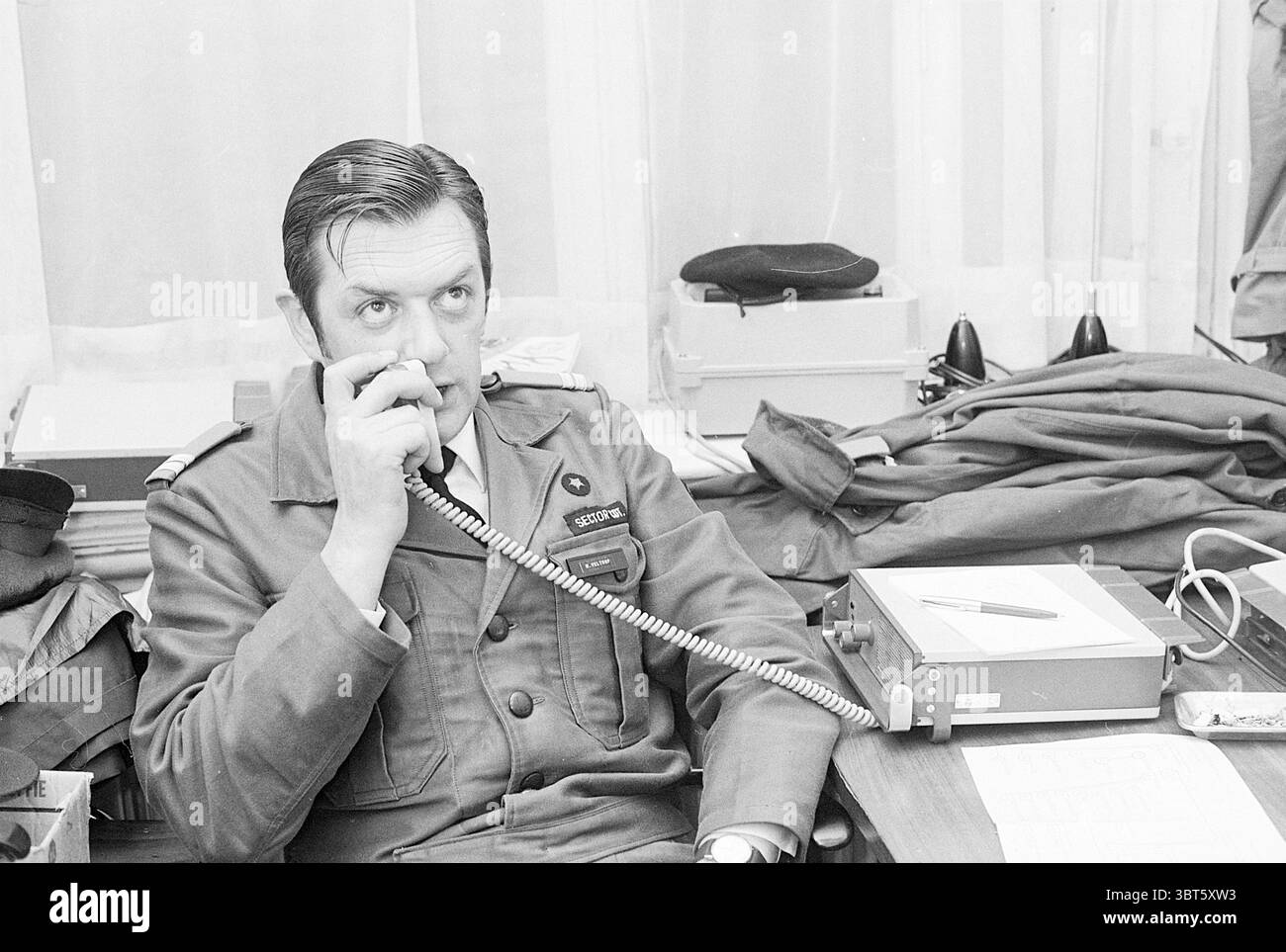 Trasferimento di grandi pensioni ospedali, Whizgle News, Dutch Desk, Paesi Bassi, 1950 - 2000 il 17-05-1971. Questi argomenti vengono visualizzati nell'immagine. In un ambiente d'ufficio vivace, un uomo in abbigliamento militare siede su una scrivania disordinata, profondamente impegnato in una conversazione attraverso un telefono. La sua uniforme è composta da un colore scuro e tenue, che suggerisce formalità e autorità. I dettagli dell'uniforme, come distintivi e insegne, suggeriscono il suo rango o ruolo in un contesto militare. La scrivania è cosparsa di vari oggetti: File, un notebook e una penna appoggiata su una superficie ben usurata. Dietro di lui, la offi Foto Stock