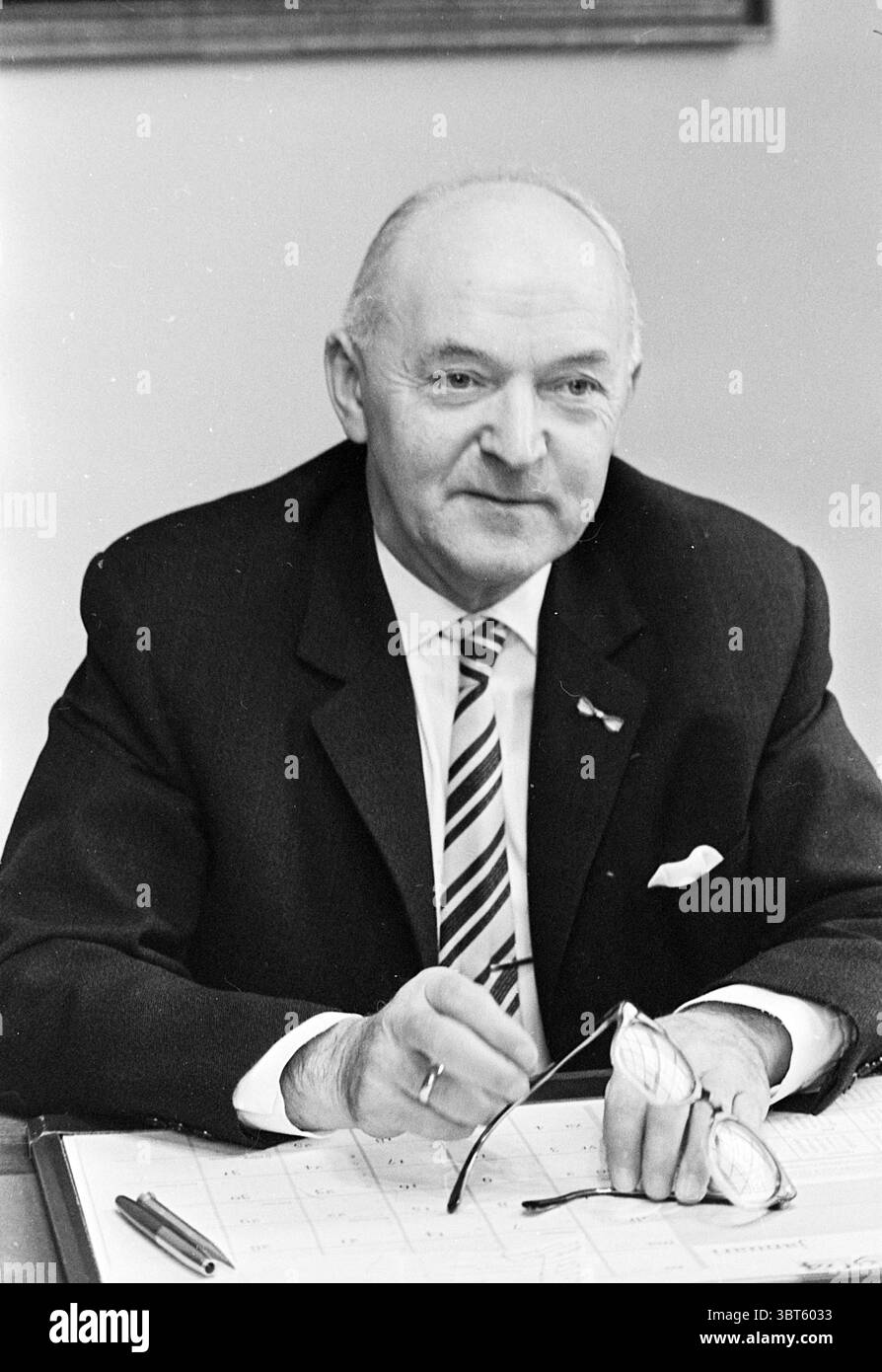 Sig. Van Wingerden segretario municipale Heemstede Persons, Whizgle News, Dutch Desk, Paesi Bassi, 1950 - 2000 il 28-01-1964. Questi argomenti vengono visualizzati nell'immagine. La scena presenta un individuo composto seduto a una scrivania. La persona è vestita con una tuta scura formale, completata da una cravatta a righe, che trasuda un aspetto professionale. Tengono un paio di occhiali delicatamente in una mano, suggerendo contemplazione o attenzione. La scrivania è adornata con vari oggetti da ufficio, tra cui una penna e quelli che possono essere documenti o un calendario, suggerendo un'area di lavoro organizzata. Lo sfondo visualizza soft, n Foto Stock