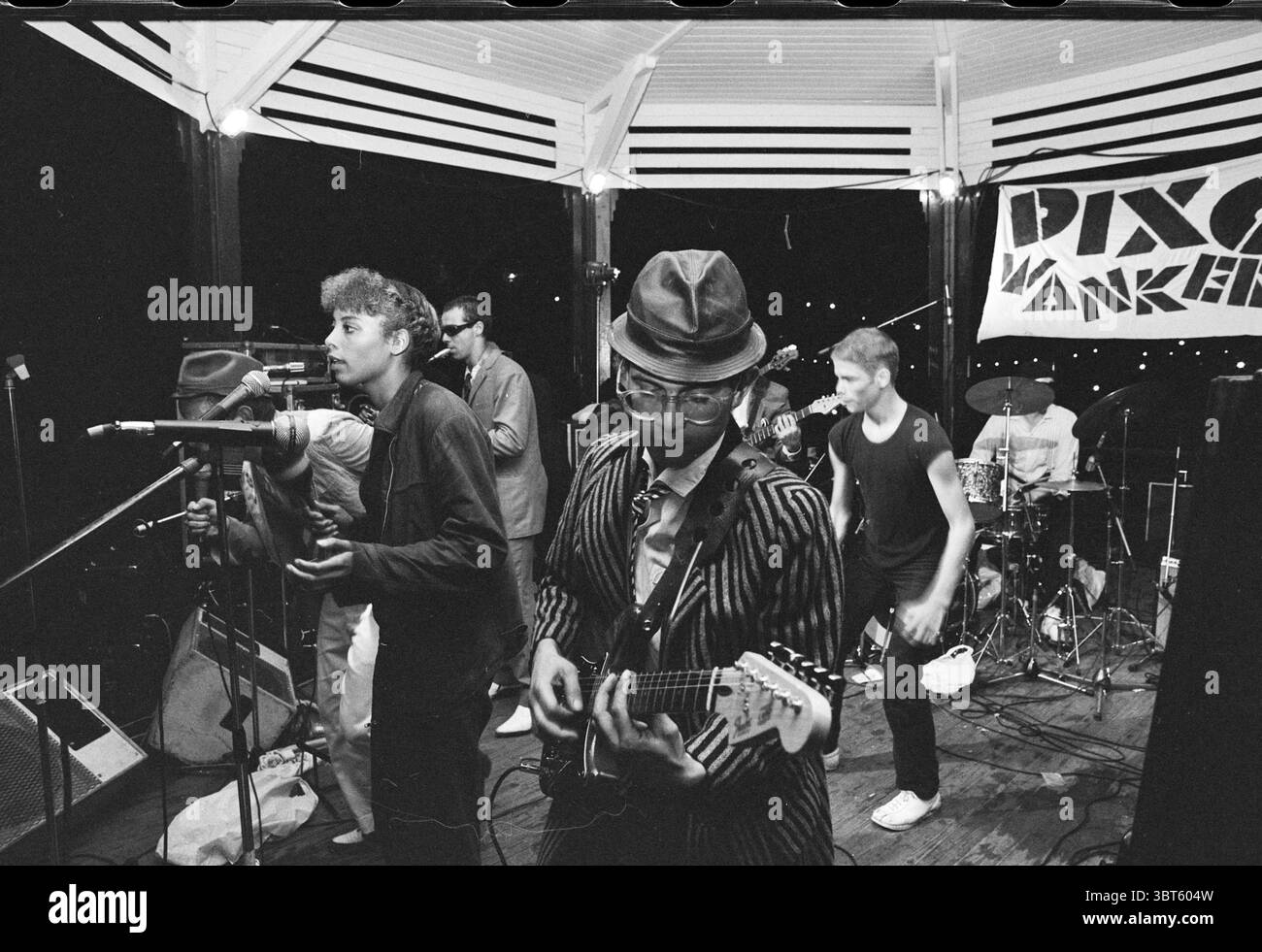 Velserbeekfestival Festival Festival festività e manifestazioni IJmuiden Velsen, Whizgle News, Dutch Desk, Paesi Bassi, 1950 - 2000 il 30-08-1980. Questi sono gli elementi dell'immagine. La scena raffigura una vivace esibizione musicale su un palcoscenico sopraelevato, caratterizzato da un'atmosfera energica. In primo piano, un gruppo diversificato di musicisti si impegna con il pubblico. Un interprete, che indossa una fedora e occhiali eleganti, si concentra sull'esecuzione di una chitarra elettrica, vestita con un'impressionante tuta a righe che contrasta con lo sfondo più scuro. Accanto a lui, un cantante, con i capelli tondi, passiona Foto Stock