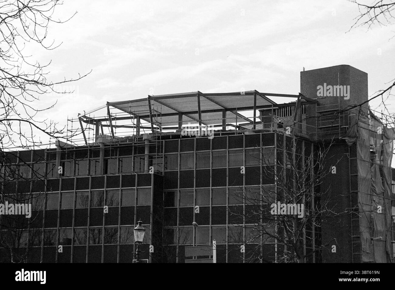 Building Social Services Zijlweg Haarlem Zijlweg, Whizgle News, Dutch Desk, Paesi Bassi, 1950 - 2000 il 02-02-1995. Questi argomenti vengono visualizzati nell'immagine. La scena presenta un edificio parzialmente costruito, che mostra la sua struttura scheletrica in modo prominente in cima. La struttura è avvolta da un mix di pannelli di vetro riflettenti e cemento scuro esposto, che contrasta nettamente con il cielo grigio chiaro. Sparsi sui livelli superiori, le travi metalliche si sporgono, suggerendo future espansioni o ristrutturazioni. L'ambiente circostante comprende rami di alberi essiccati che si estendono dagli angoli, A. Foto Stock