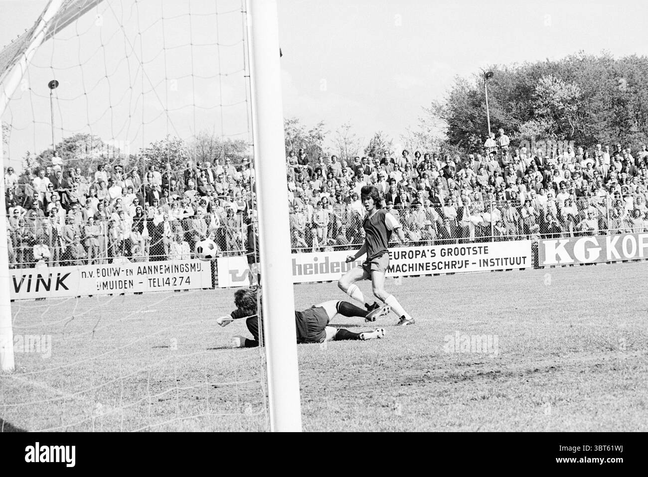 Partita di calcio., Whizgle News, Dutch Desk, Paesi Bassi, 1950 - 2000. Questi sono gli argomenti nell'immagine. In una vivace scena sportiva all'aperto, l'attenzione è concentrata su una partita di calcio che si svolge sotto un cielo azzurro. L'atmosfera è elettrica, piena di emozioni mentre gli spettatori riempiono le tribune, i loro volti animati da applausi e anticipazioni. Il campo erboso è ben tenuto e verde vivace, in contrasto con i toni più scuri del vicino porticciolo. Un giocatore con una maglia a strisce si trova nel bel mezzo di un gioco dinamico, manovrando abilmente la palla verso la porta. La sua postura è intensa e foc Foto Stock