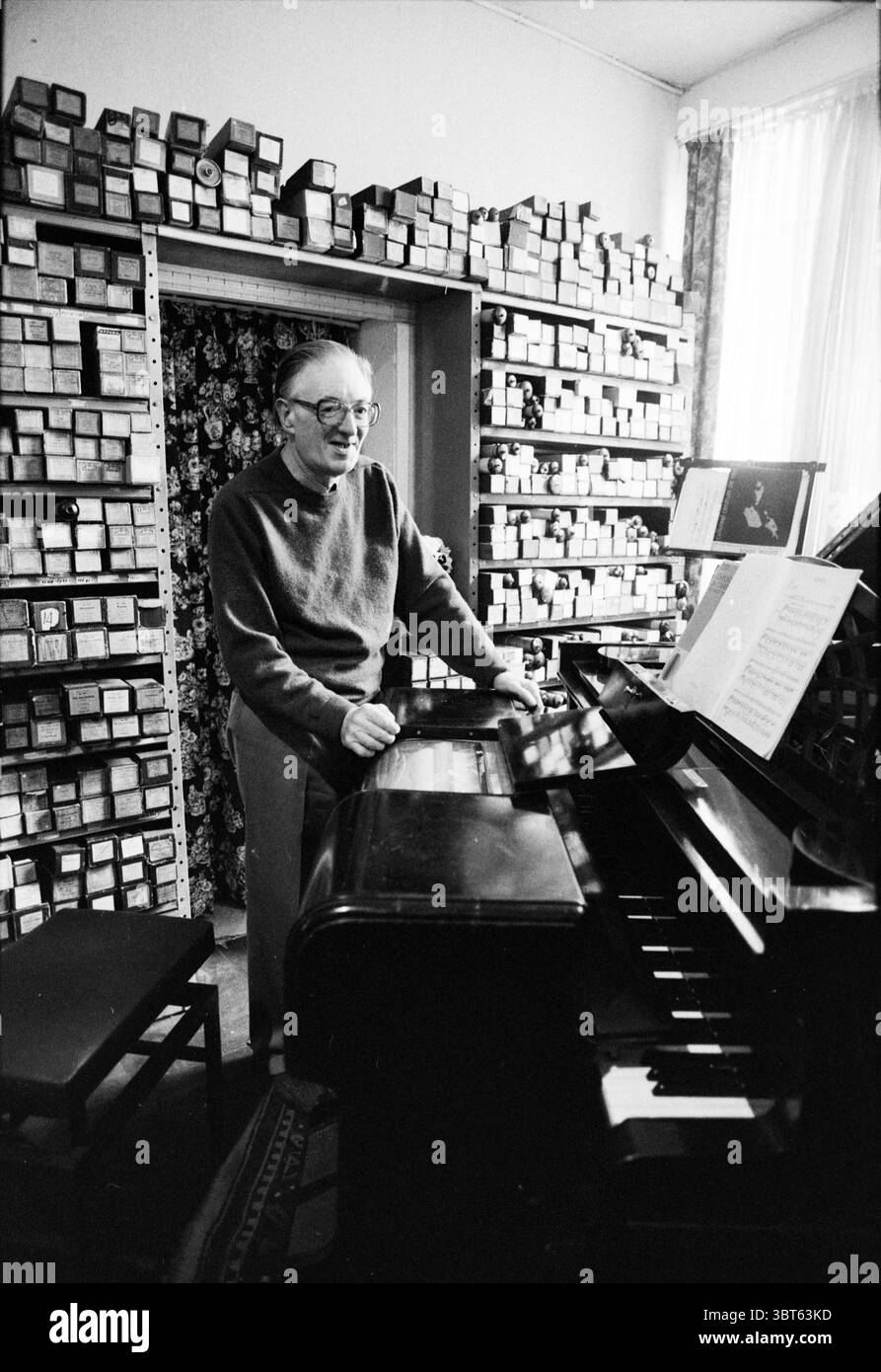 Groos + pianola, Whizgle News, Dutch Desk, Paesi Bassi, 1950 - 2000 il 14-10-1991. Questi argomenti vengono visualizzati nell'immagine. In una camera accogliente e un po' disordinata, un anziano si erge accanto a un pianoforte nero lucido. Indossa un caldo maglione oversize che suggerisce comfort e familiarità. I suoi occhiali poggiano sul naso, incorniciando un'espressione allegra che rivela un senso di gioia nell'ambiente circostante. Lo sfondo è pieno di ripiani che si estendono dal pavimento al soffitto, strettamente pieni di numerose scatole, il loro contenuto lascia intendere spartiti musicali o ricordi, probabilmente riflettendo anni di dedica Foto Stock