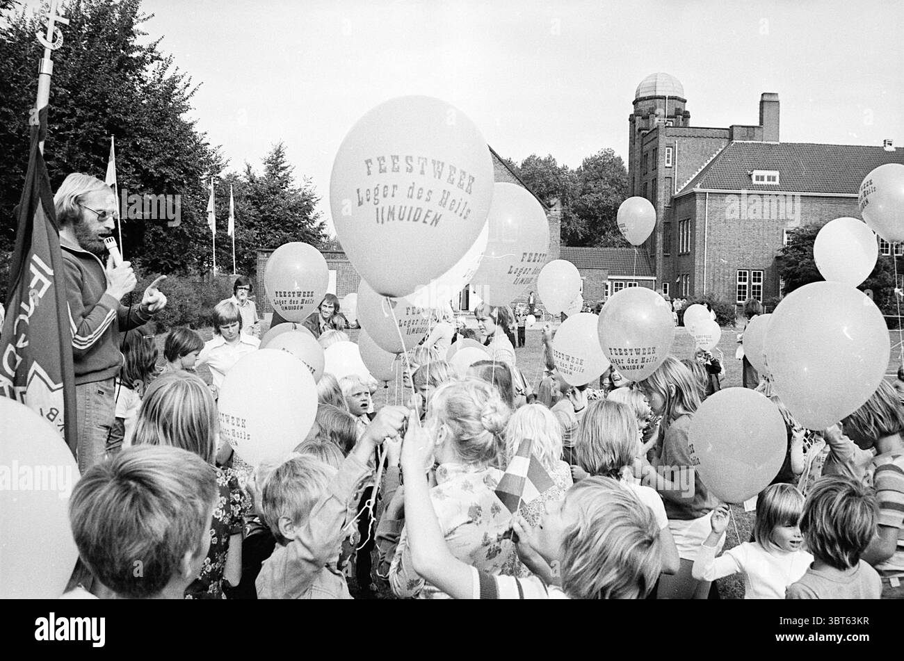 Settimana di festa IJmuiden, Whizgle News, Dutch Desk, Paesi Bassi, 1950 - 2000 nel 1975. Questi sono gli argomenti nell'immagine. La scena cattura un energico raduno all'aperto, probabilmente una festa o un festival, caratterizzato da un'atmosfera vivace. Una folla di bambini, vestiti con abiti colorati, è gioiosamente coinvolgente l'uno con l'altro. Il loro abbigliamento presenta motivi vivaci che aggiungono un tono allegro alla scena, mentre alcuni bambini tengono in mano palloncini di varie dimensioni. I palloncini sono disponibili in morbide tonalità pastello, contribuendo all'atmosfera festosa. In primo piano, un oratore, probabilmente un maschio adulto, stan Foto Stock
