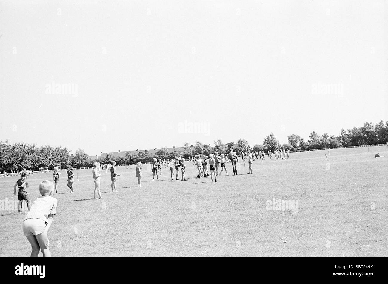 Giornata dello sport per bambini, Whizgle News, Dutch Desk, Paesi Bassi, 1950 - 2000 il 15-07-1969. Questi argomenti vengono visualizzati nell'immagine. La scena cattura un ambiente all'aperto vivace, probabilmente una giornata estiva, caratterizzato da una luce solare intensa che proietta un forte contrasto nel paesaggio. Il primo piano presenta una distesa erbosa, lussureggiante e ben tenuto, dove numerosi bambini sono impegnati in varie attività. Nel terreno di mezzo, gruppi di bambini, vestiti con abiti estivi leggeri, sono sparsi in tutta l'area erbosa. Il loro abbigliamento è prevalentemente di colore chiaro, con un mix di pantaloncini e t-shirt, W Foto Stock