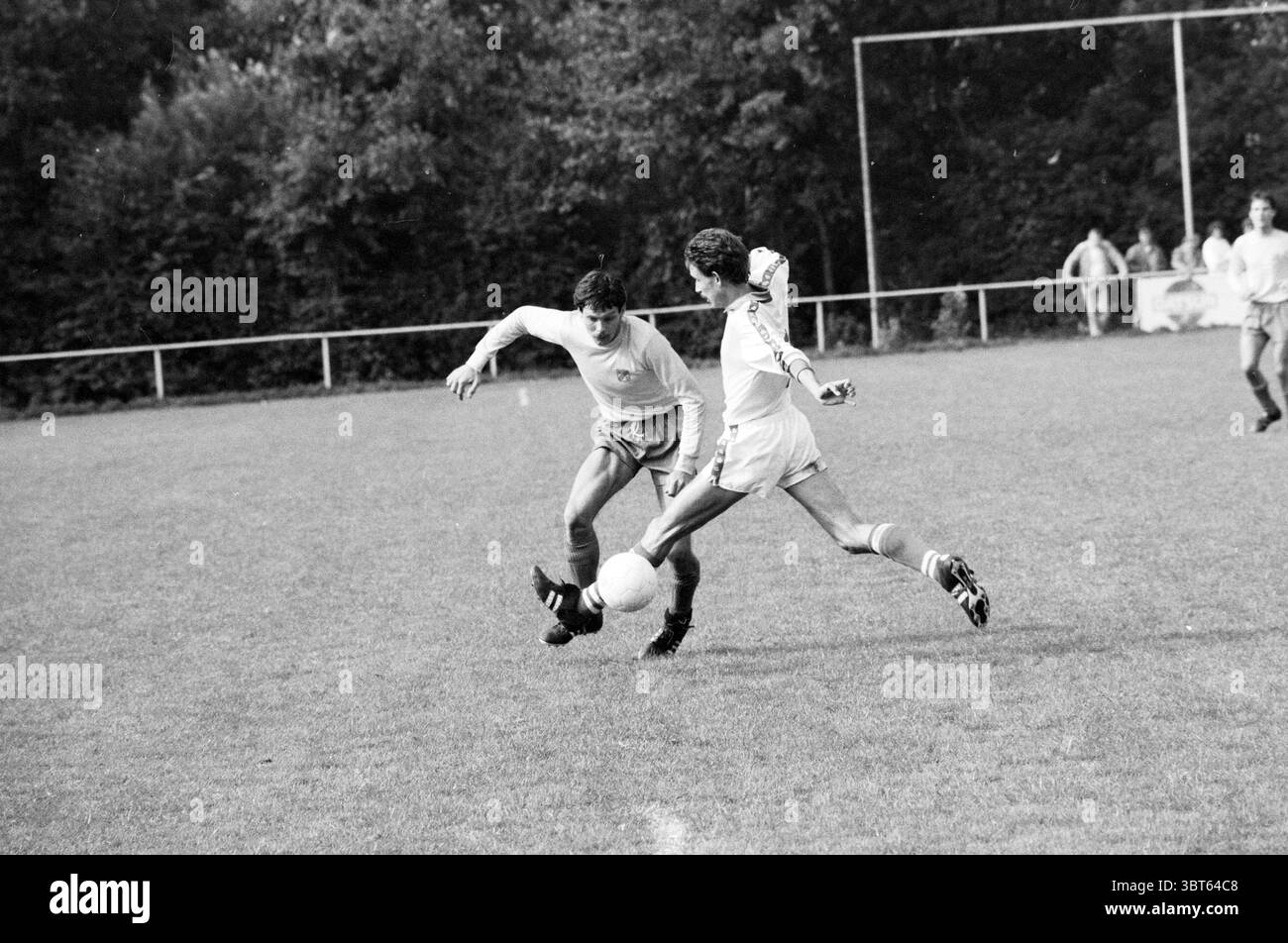 ODIN - ZOB Football Odin, Whizgle News, Dutch Desk, Paesi Bassi, 1950 - 2000 il 13-09-1986. Questi argomenti sono mostrati nell'immagine. La scena raffigura un momento intenso in una partita di calcio ambientata su un campo erboso circondato da un ambiente rustico di alberi in varie sfumature di verde. I giocatori, vestiti con divise a contrasto, una con un top leggero con pantaloncini corti e l'altra con un outfit più scuro, sono impegnati in un gioco dinamico. Il giocatore in bianco si inclina in avanti, manovrando abilmente la palla lontano dall'avversario, che sembra essere affondato verso di lui con determinazione concentrata. Il campo è vivace, Foto Stock