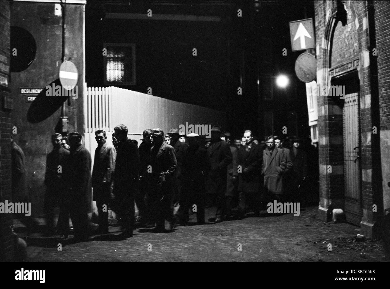 R.K.Stille Omgang Noord Zuid Hollandse Vervoersmaatschappij N.V. NZ, Whizgle News, Dutch Desk, Paesi Bassi, 1950 - 2000 il 16-03-1963. Questi argomenti vengono visualizzati nell'immagine. La scena trasmette un'atmosfera cupa e misteriosa, illuminata da fonti luminose poco chiare e sparse. Una fila di persone, rivestite da lunghi soprabiti e cappelli, si muove in un unico file lungo uno stretto vicolo acciottolato. Le loro espressioni sono serie e contemplative, aggiungendo alla gravità complessiva del momento. Lo sfondo presenta pareti in mattoni, alcune adornate con forme vaghe e ombre, che creano un senso di profondità e di recinzione. A. Foto Stock