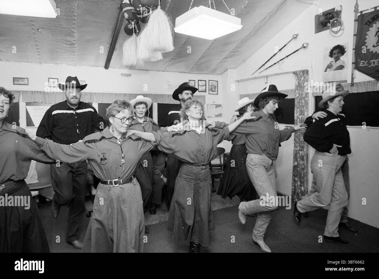 Danza country Vijfhuizen Vijfhuizen, Whizgle News, Dutch Desk, Paesi Bassi, 1950 - 2000 il 28-01-1992. L'immagine mostra questi argomenti. La scena cattura un vivace raduno al coperto, probabilmente ambientato in una sala rustica adornata con arredamento a tema occidentale. La tavolozza dei colori presenta principalmente toni tenui di marrone, grigio e blu scuro, evocando un senso di nostalgia e calore. Un gruppo di individui, un mix di uomini e donne, sono impegnati in una danza coordinata di linea. Le loro espressioni allegre e le pose animate suggeriscono uno spirito di cameratismo e divertimento. La maggior parte indossa cappelli da cowboy, che si aggiungono all'ambi occidentale Foto Stock