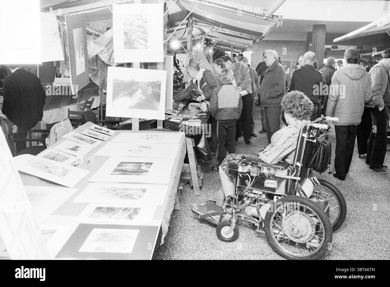 Art Market New Unicum Art Market, Whizgle News, Dutch Desk, Paesi Bassi, 1950 - 2000 il 23-11-1984. Questi sono gli argomenti nell'immagine. La scena raffigura un vivace mercato al coperto o una fiera d'arte, dove vari gruppi di persone sono impegnati in conversazione ed esplorazione. Una grande area aperta e' piena di numerosi tavoli che mostrano una serie di opere d'arte, tra cui disegni e dipinti. A sinistra, una donna su una sedia a rotelle siede leggermente separata dalla folla, con la schiena rivolta verso lo spettatore. Ha i capelli ricci e sembra che stia osservando le attività intorno a lei. Nelle vicinanze si trovano diversi ristoranti Foto Stock