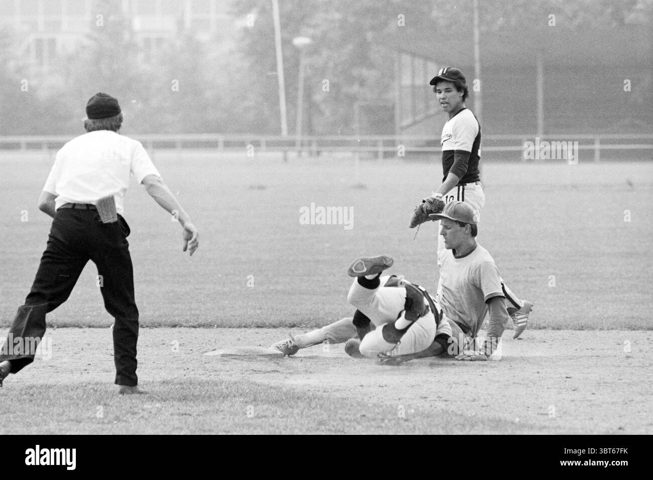 Rooswijk- Black Lions Baseball Rooswijk, Whizgle News, Dutch Desk, Paesi Bassi, 1950 - 2000 il 31-07-1982. Questi argomenti sono mostrati nell'immagine. In una scena sportiva dinamica, due giocatori sono impegnati in un momento di tensione su un diamante da baseball. Il giocatore che scivola nella base viene catturato a metà movimento, con le gambe allungate e un braccio allungato, suggerendo un momento di urgenza e competitività. Indossa una maglia di colore chiaro che contrasta nettamente con lo sporco marrone della base. Di fronte a lui, un fielder è pronto alla base. Questo giocatore, che indossa un'uniforme più scura, è pronto e concentrato, demoni Foto Stock