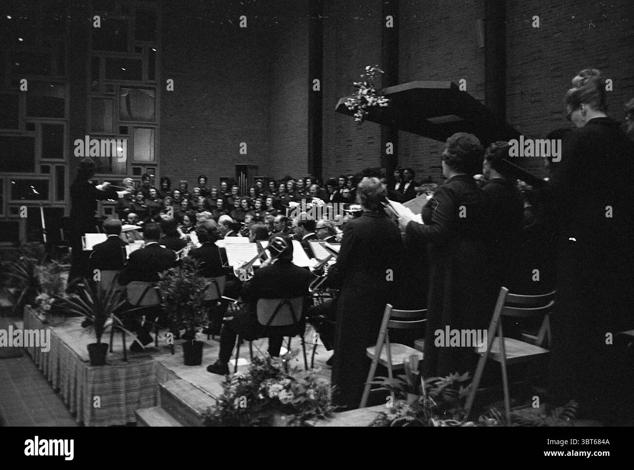 Concerto di coro e orchestra in chiesa., Whizgle News, Dutch Desk, Paesi Bassi, 1950 - 2000. Questi sono gli argomenti nell'immagine. La scena si sviluppa in una grande sala poco illuminata, che esalta un'atmosfera cupa e allo stesso tempo celebrativa. Un grande gruppo di musicisti e cantanti riempie lo spazio, creando un ricco arazzo di suoni. In primo piano, un gruppo eterogeneo di musicisti occupa file di semplici sedie di metallo, concentrandosi intensamente sui loro spartiti musicali. Gli strumenti in ottone brillano sottilmente sotto la luce scarsa, mentre i venti e le corde creano un mix invitante e armonioso. Il loro abbigliamento, per lo più in buio h Foto Stock