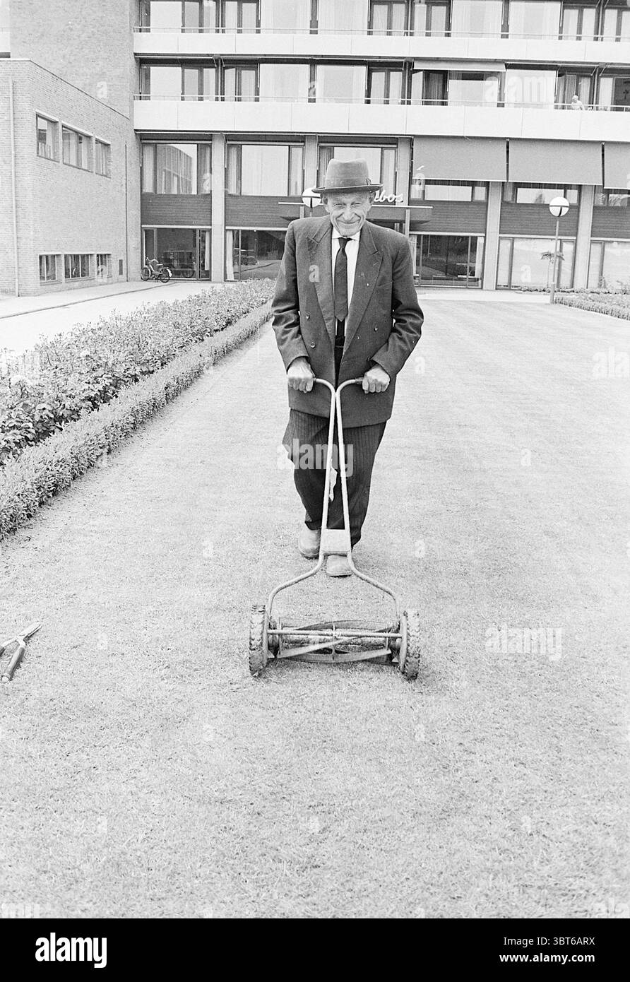 90 y. giardiniere di Overbos Persons, Whizgle News, Dutch Desk, Paesi Bassi, 1950 - 2000 il 14-07-1969. Questi sono gli elementi dell'immagine. La scena cattura un uomo anziano che spinge diligentemente un rasaerba vintage manuale su una distesa di erba appena tagliata. È vestito con una tuta scura e intelligente con righe e un cappello fedora abbinato, suggerendo un senso di formalità e forse un'epoca passata. L'erba sotto i suoi piedi è un verde uniforme e vibrante, che contrasta splendidamente con le linee nitide del rasaerba, che ha un aspetto metallico e resistente agli agenti atmosferici, evocando un senso di nostalgia. Surround Foto Stock